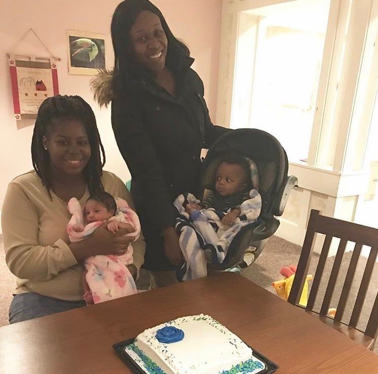 Woman with two babies at a dining table beside a birthday cake, smiling indoors.