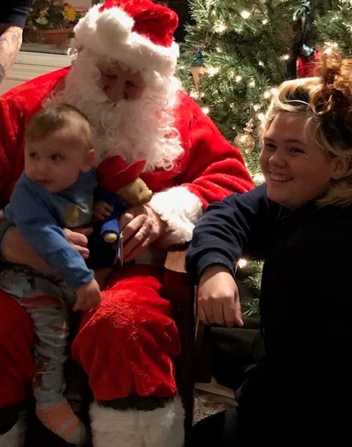 Santa holding a toddler beside a smiling woman in front of a lit Christmas tree