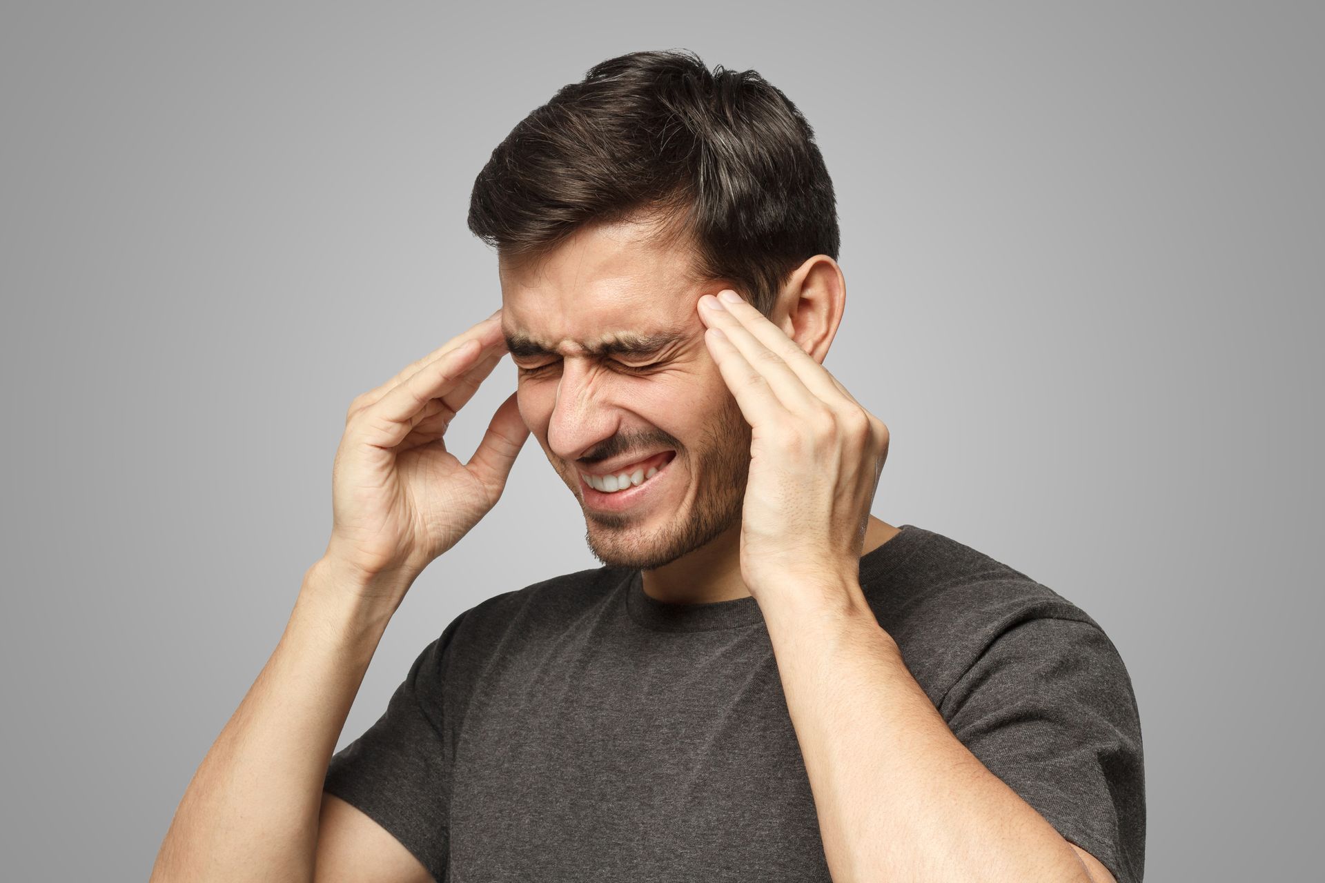 Homem com os olhos fechados, fazendo careta, segurando as têmporas, sugerindo dor de cabeça, contra um fundo cinza.