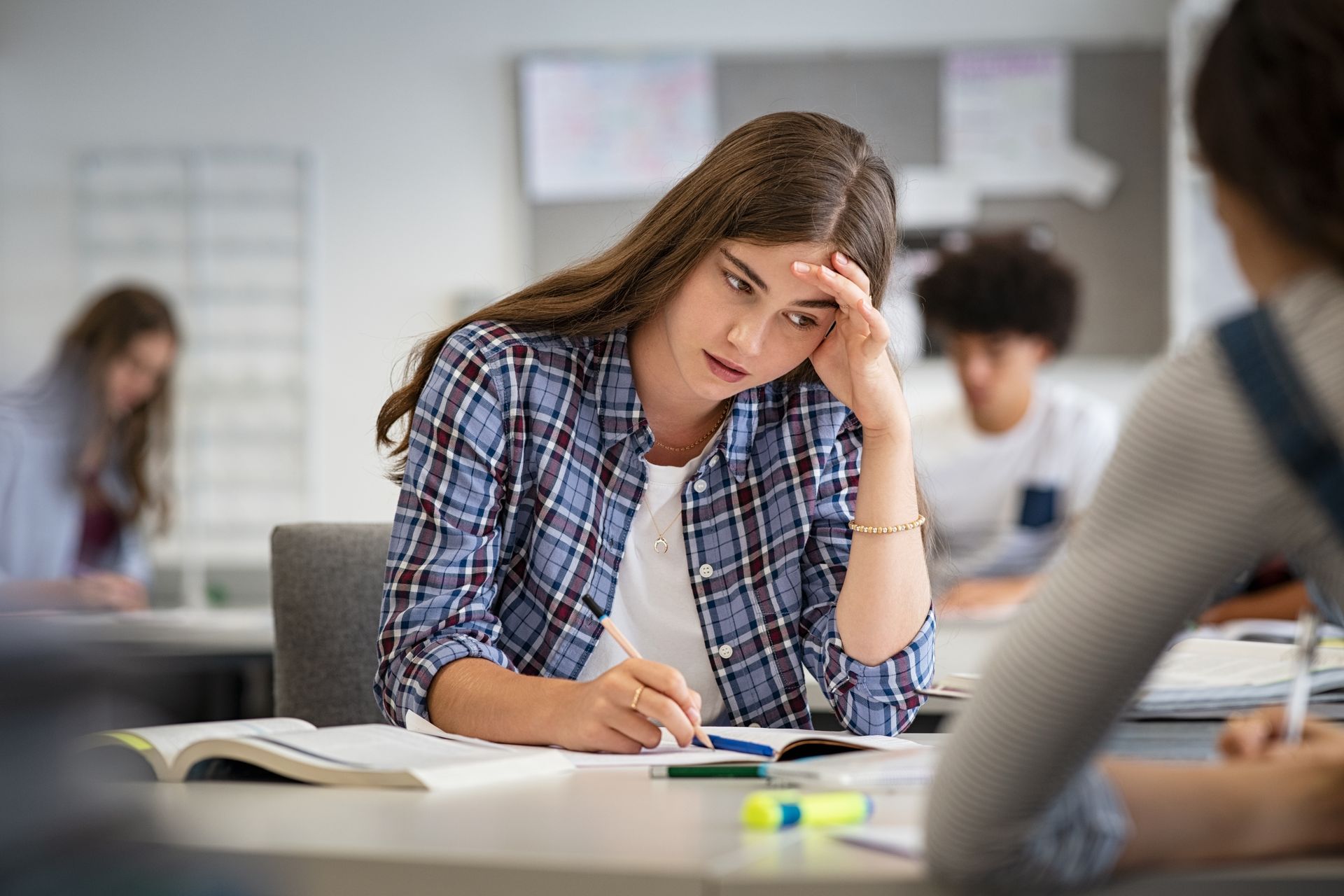Student in plaid shirt, furrowed brow, writing in notebook at desk in classroom.