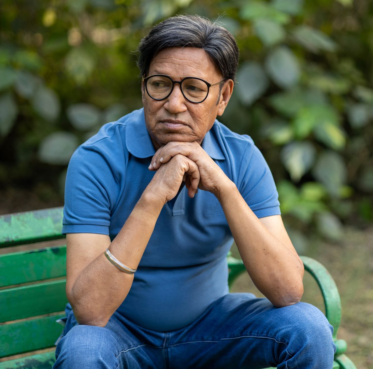 Man in glasses sits on park bench, hands under chin, deep in thought. Blue shirt, bracelet.