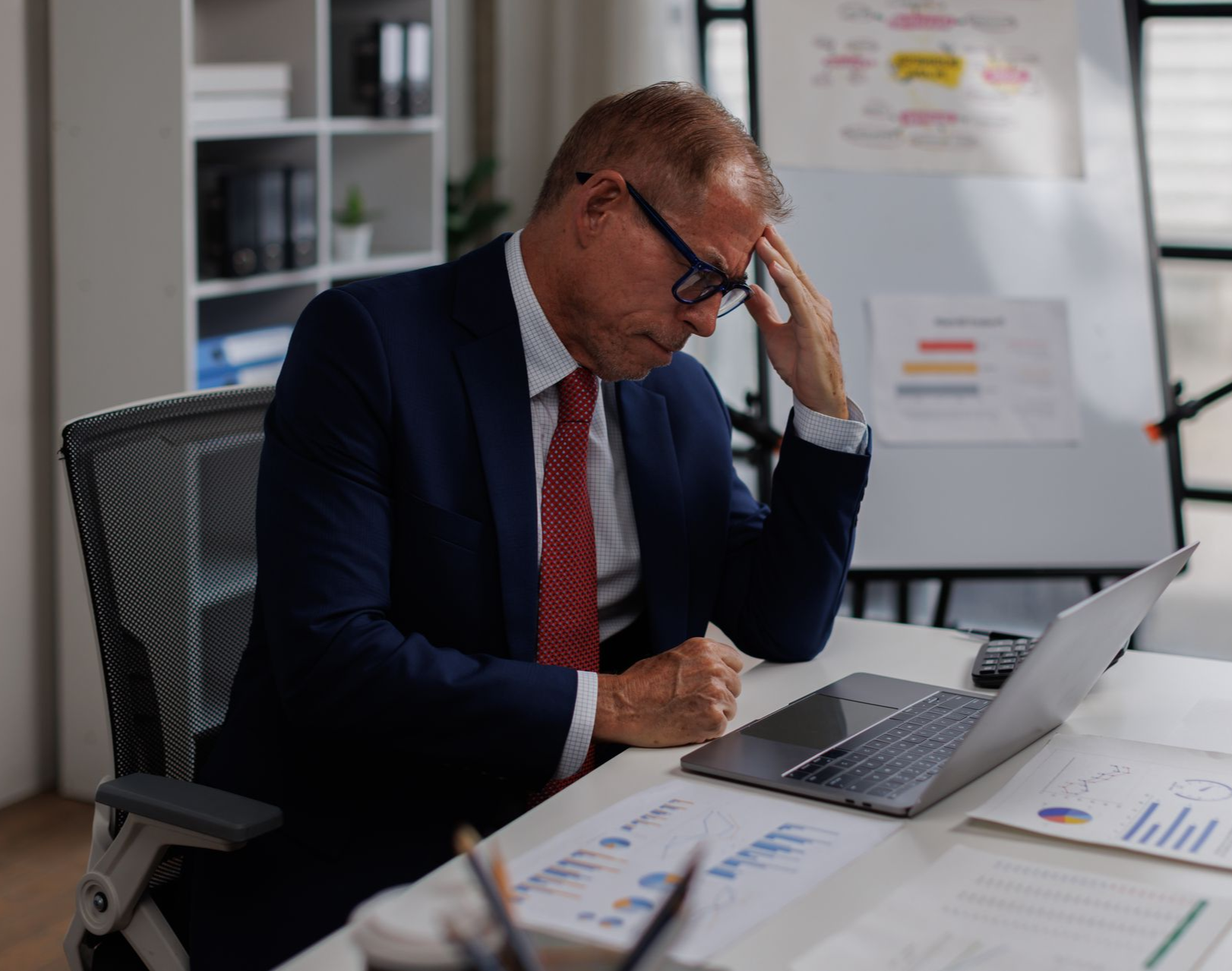 Man in suit at desk, stressed, hand on head, looking at laptop and papers.