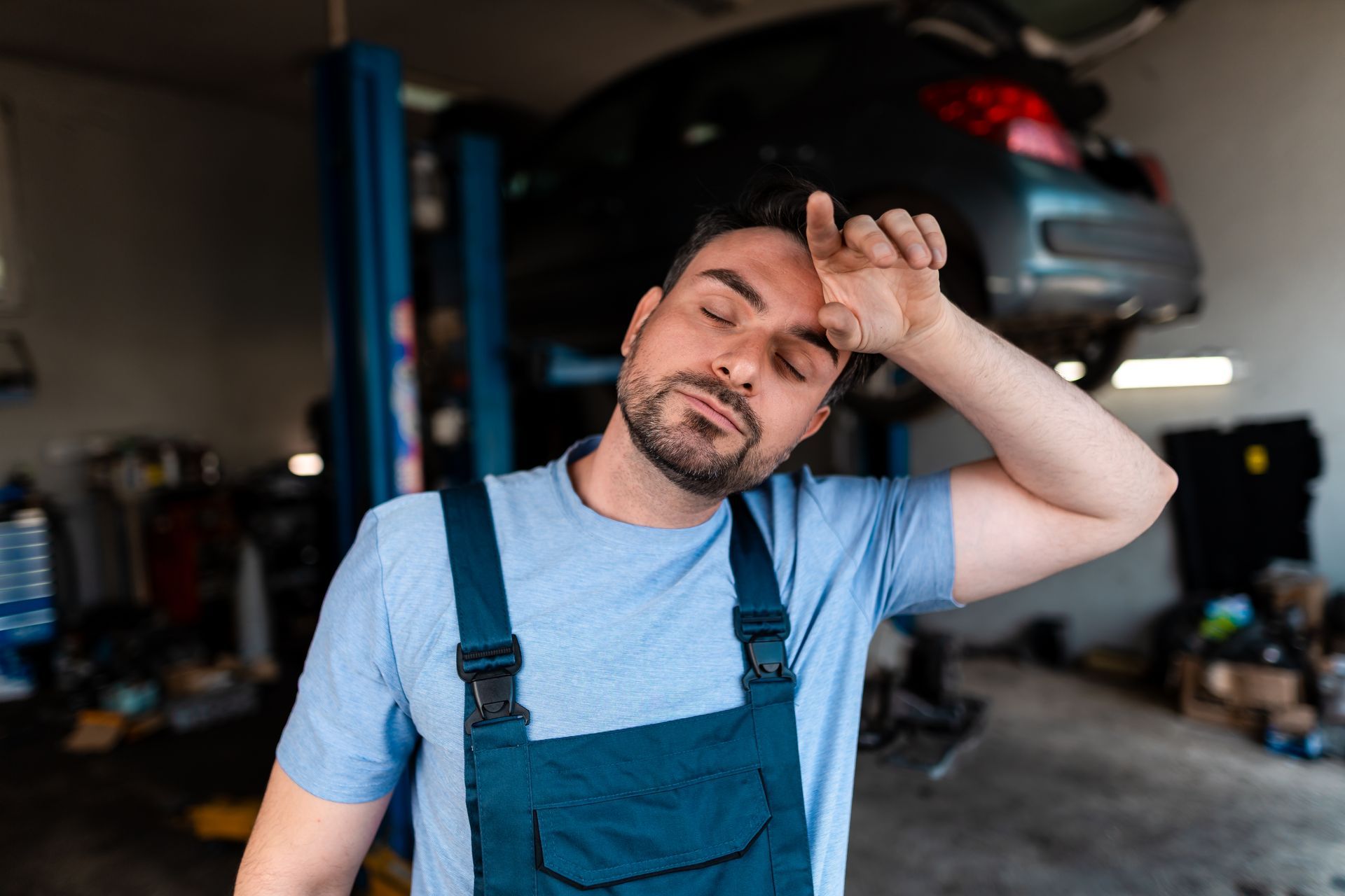 Mechanic wiping sweat from brow, eyes closed, in a car repair shop with a car on a lift.