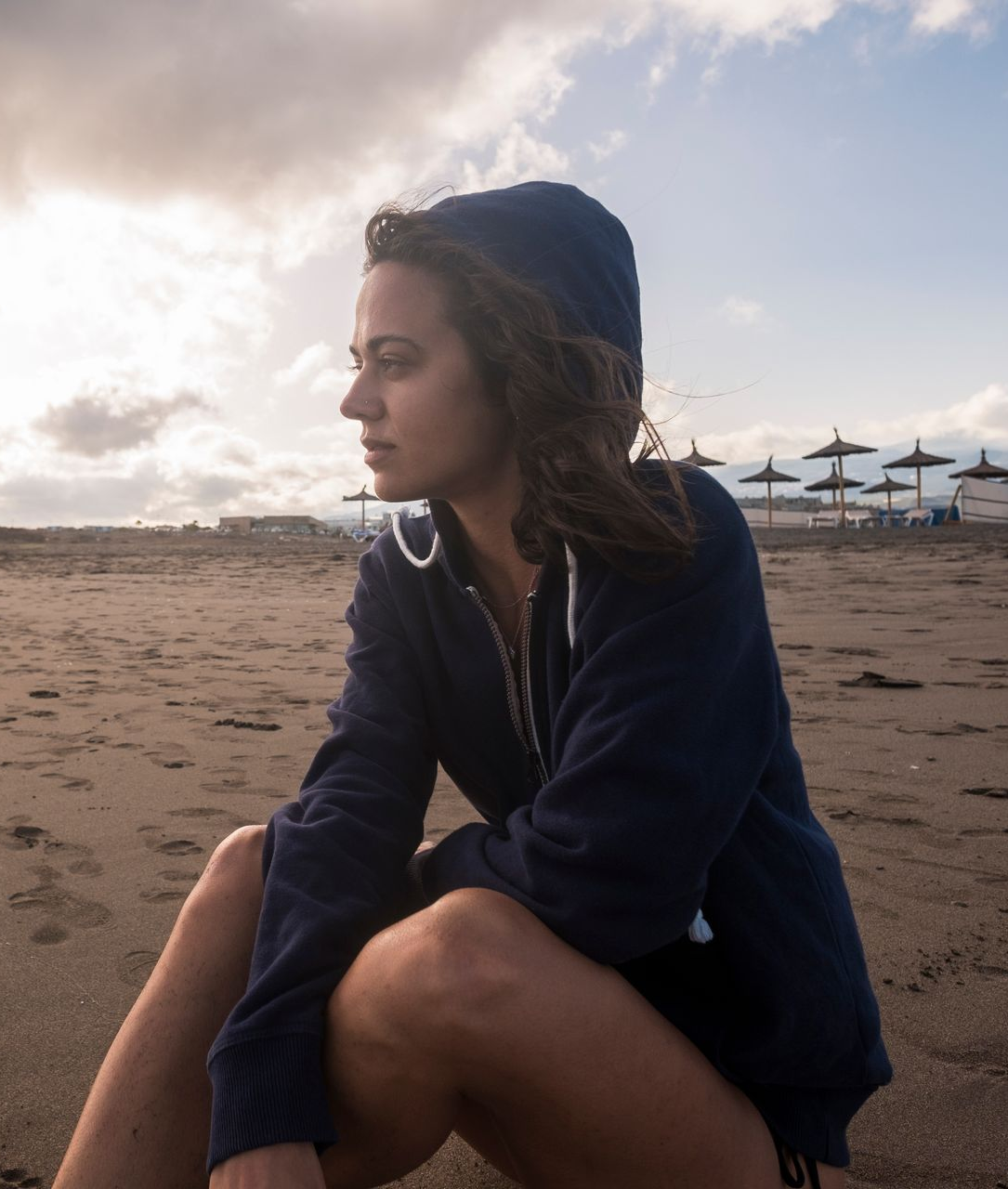 Woman in blue hoodie sits on beach, looking away, cloudy sky.