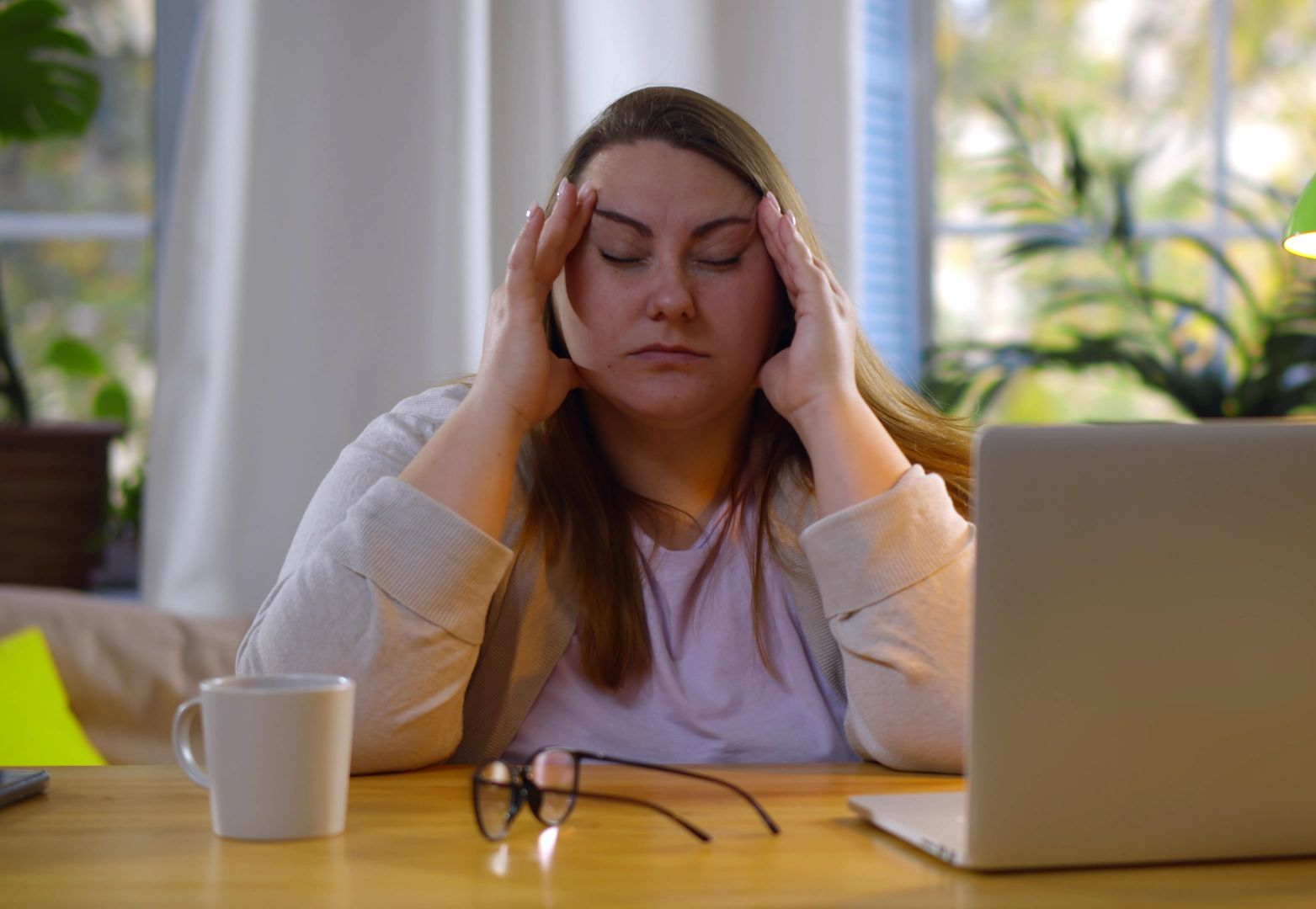 Woman with closed eyes, hands on head, at desk with laptop, glasses, and mug.