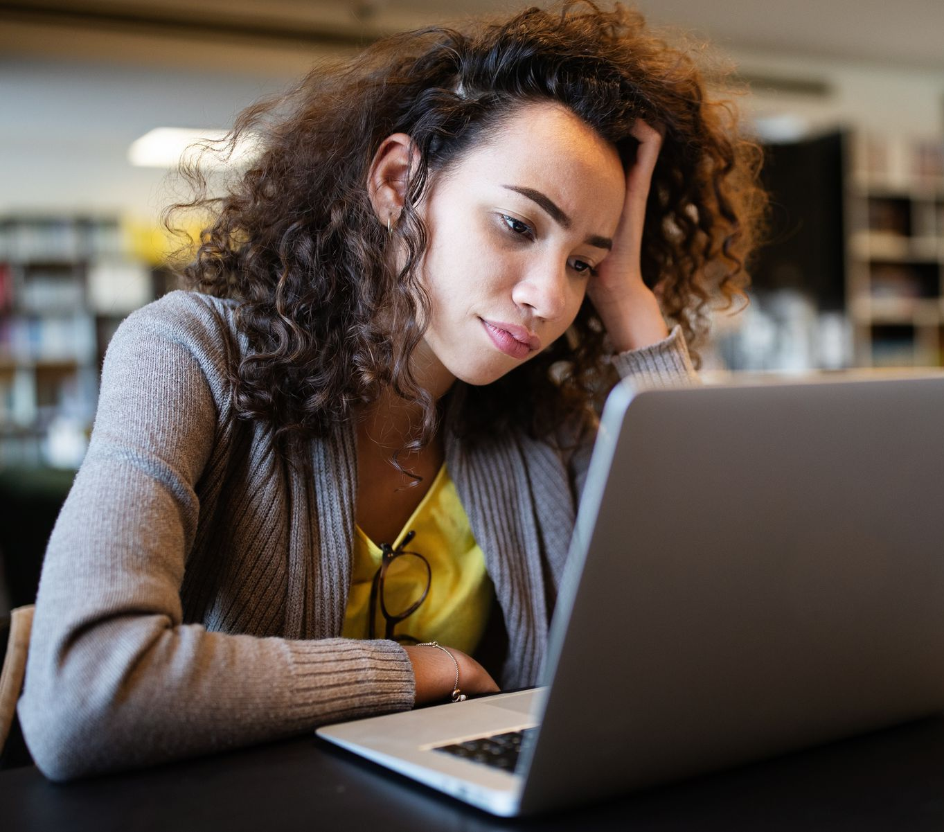 Woman at a laptop looks stressed, resting her head on her hand. In a library, with bookshelves in the background.