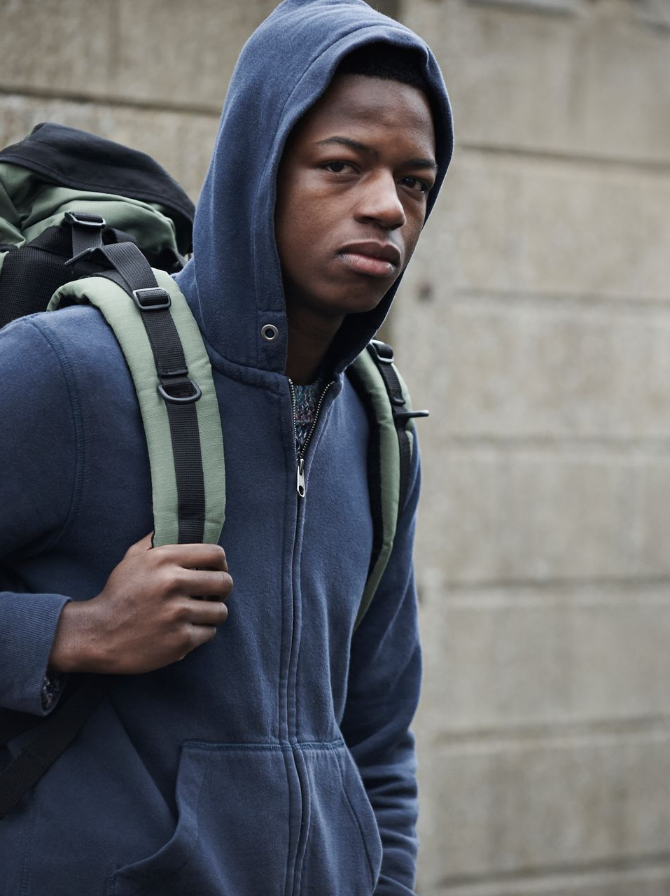 Person in a blue hooded sweatshirt and backpack, stands near a concrete wall, looking towards camera.