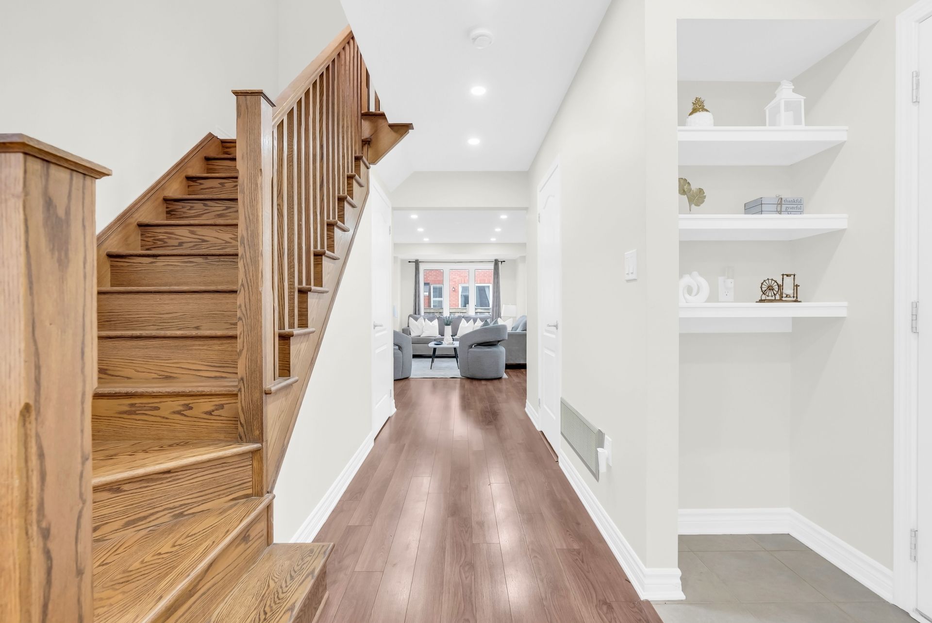 Hallway with wood staircase, white walls, built-in shelves, and hardwood floor leading to a living room.