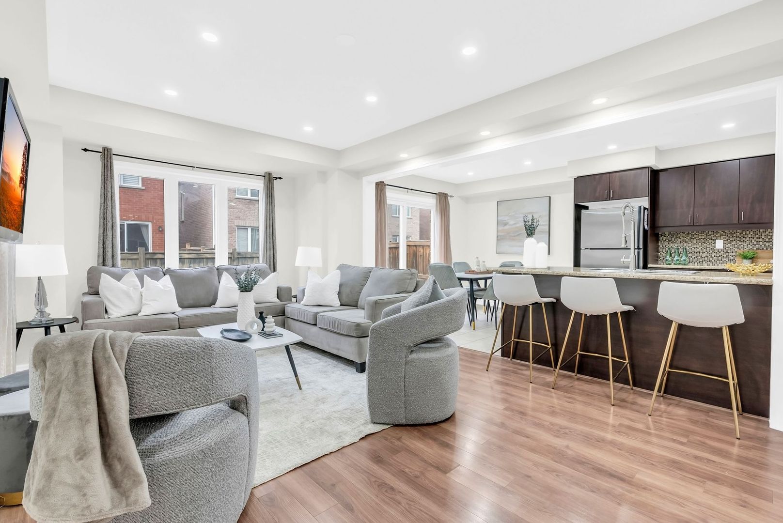 Bright open living room and kitchen with gray furniture, wood floors, and white bar stools