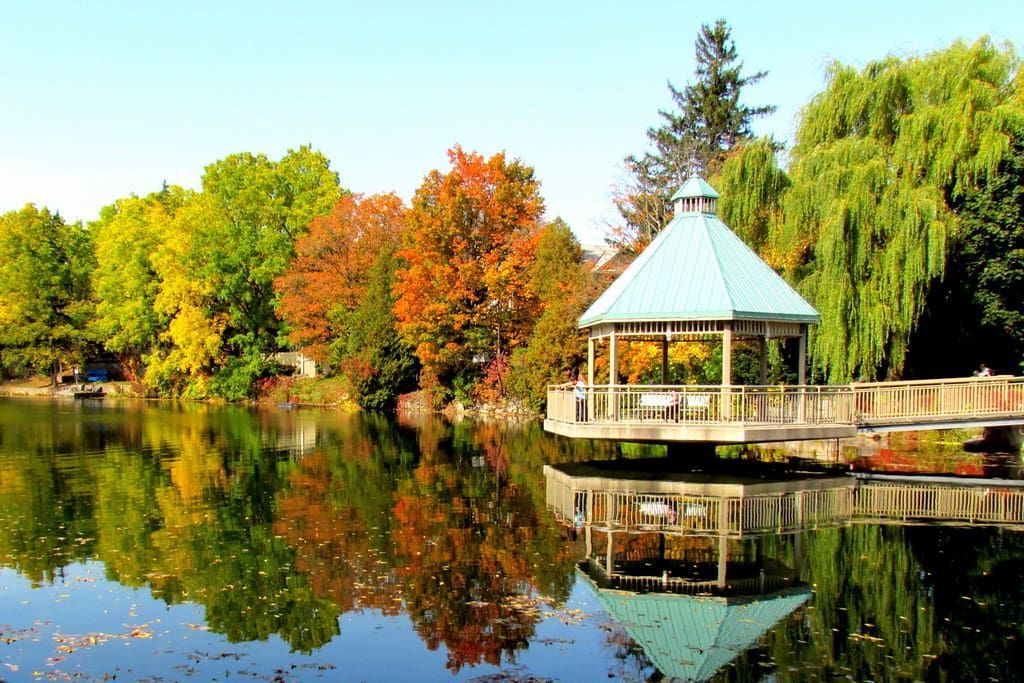 Autumn trees reflect on a calm lake beside a small gazebo with a blue roof