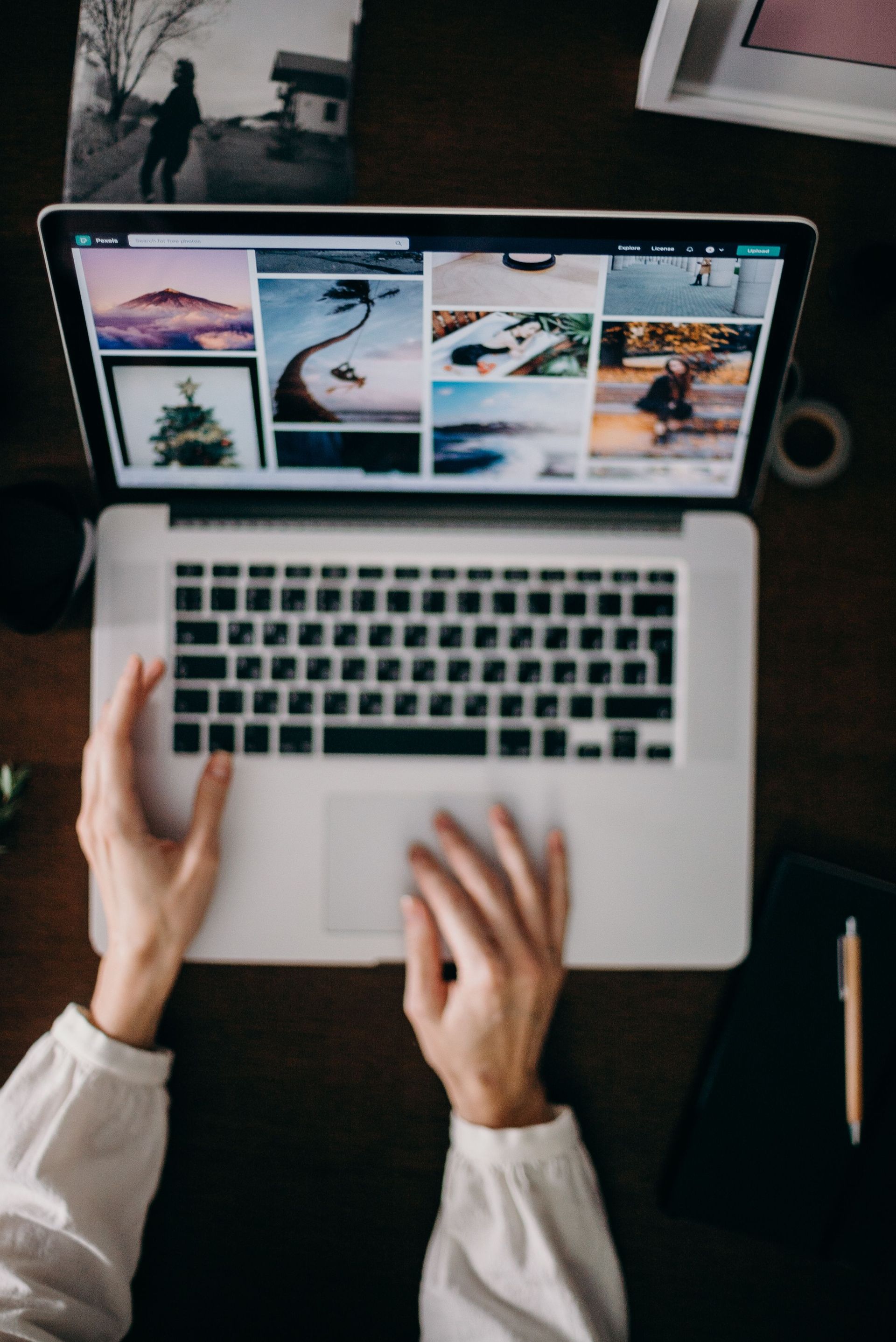 Hands typing on a laptop displaying a photo grid on a dark desk with a pencil nearby