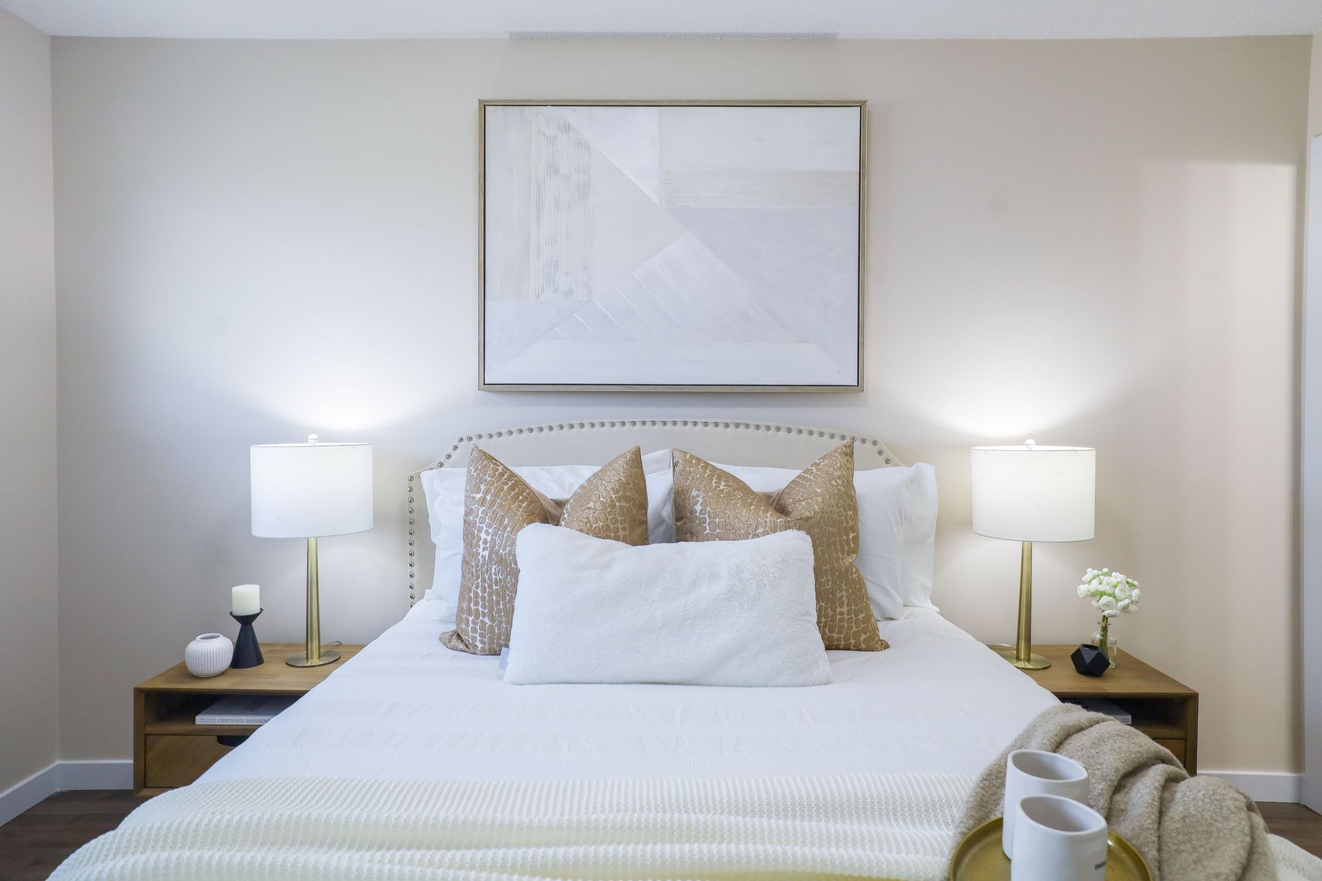 Neatly made bed with white bedding, matching lamps on nightstands, and a framed abstract print above the headboard.