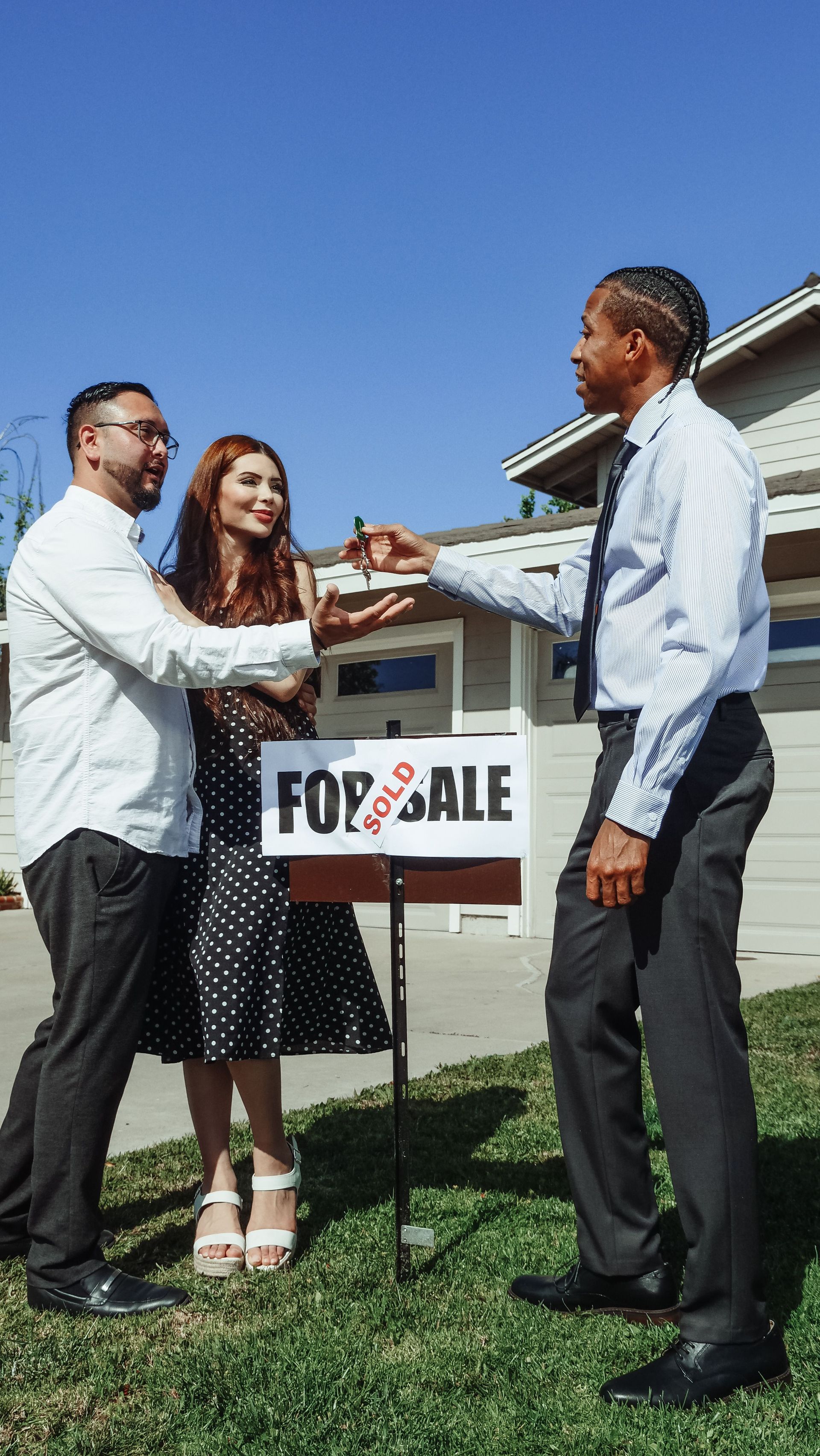 Three people shake hands beside a for sale sign in front of a house under a clear blue sky.