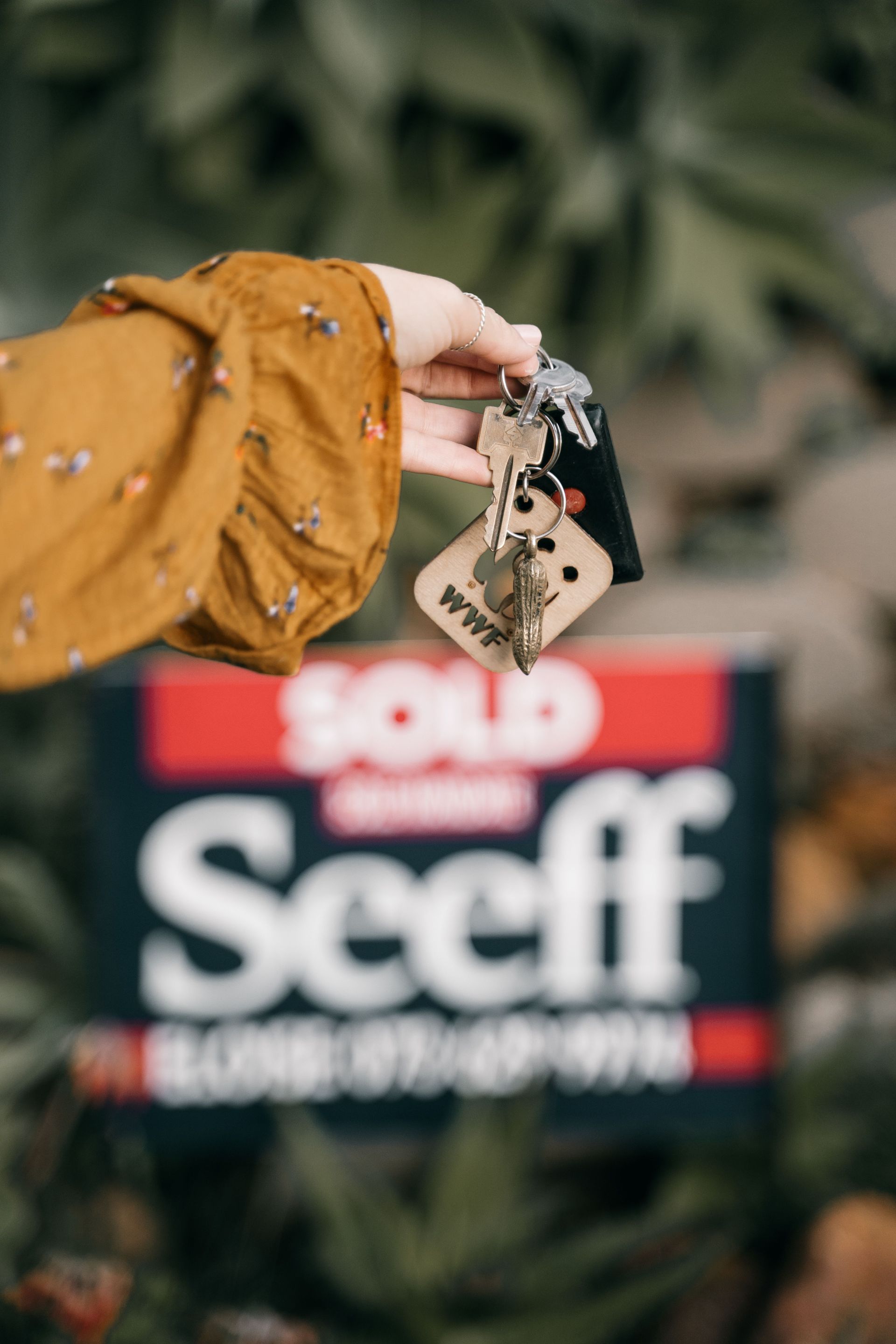 Hand holding car keys in front of a blurred red and black “Sold” real estate sign