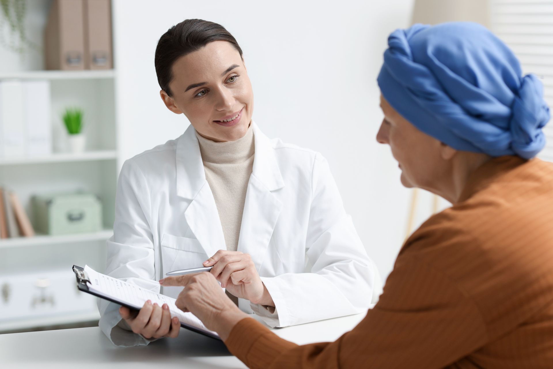 Doctor consulting with a patient in a clinic, holding a clipboard and speaking across a desk.