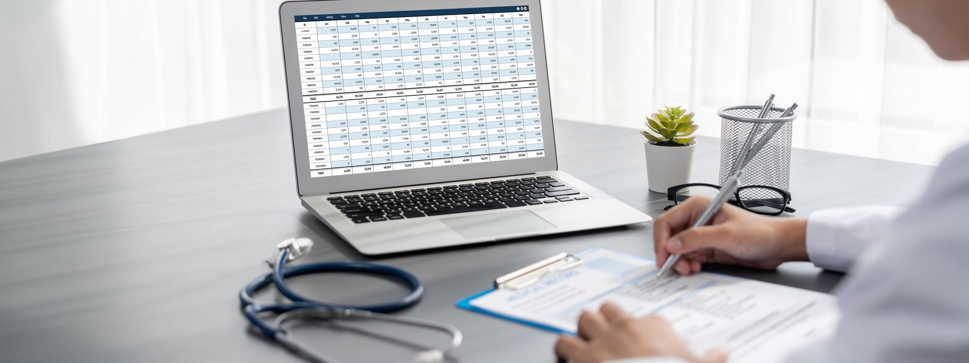 Laptop displaying charts beside a hand taking notes at a desk with stethoscope and coffee cup