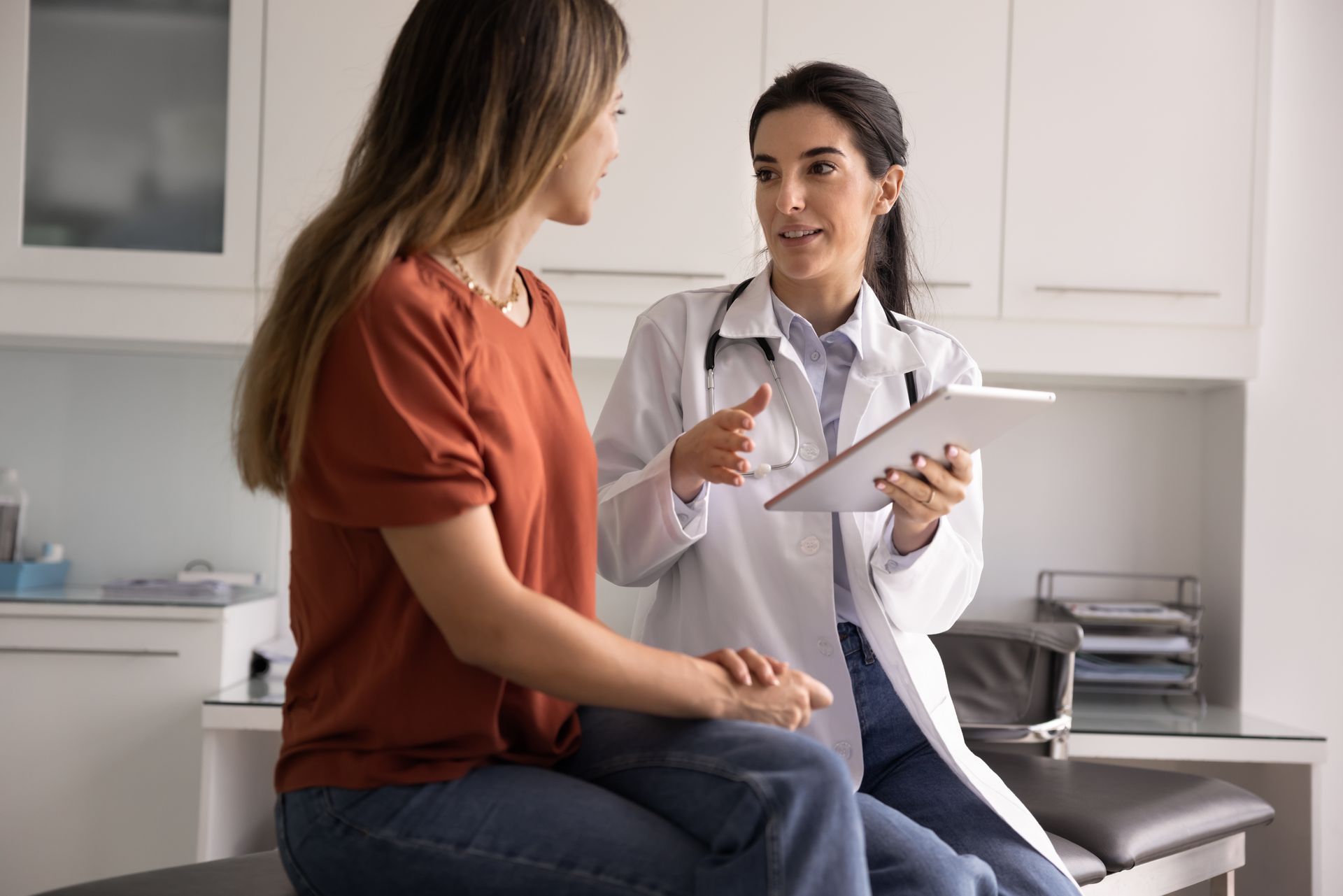 Doctor speaking with a seated patient in a bright clinic, holding a tablet during a consultation.