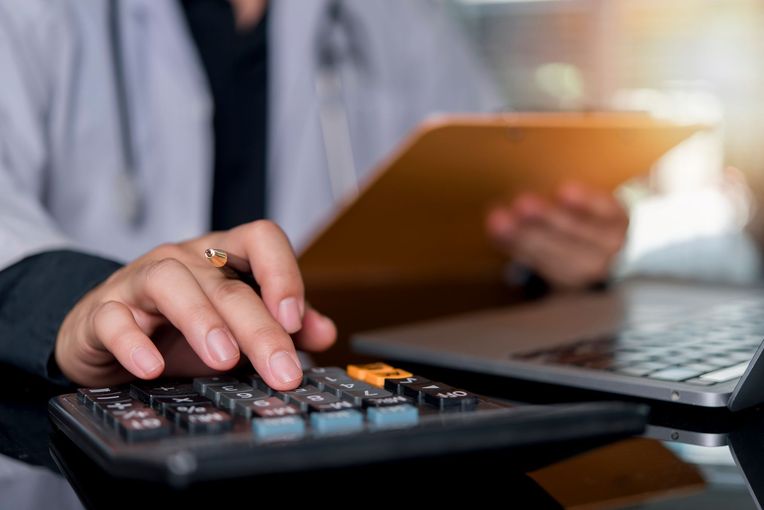 Hand using calculator beside laptop on a desk, with a clipboard in the background