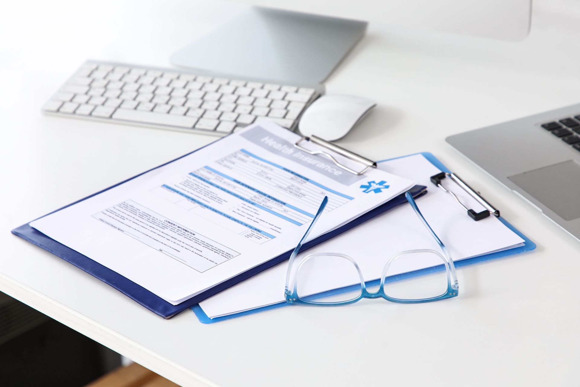 Clipboard with documents, blue eyeglasses, and pen on a white office desk with keyboard and laptop.
