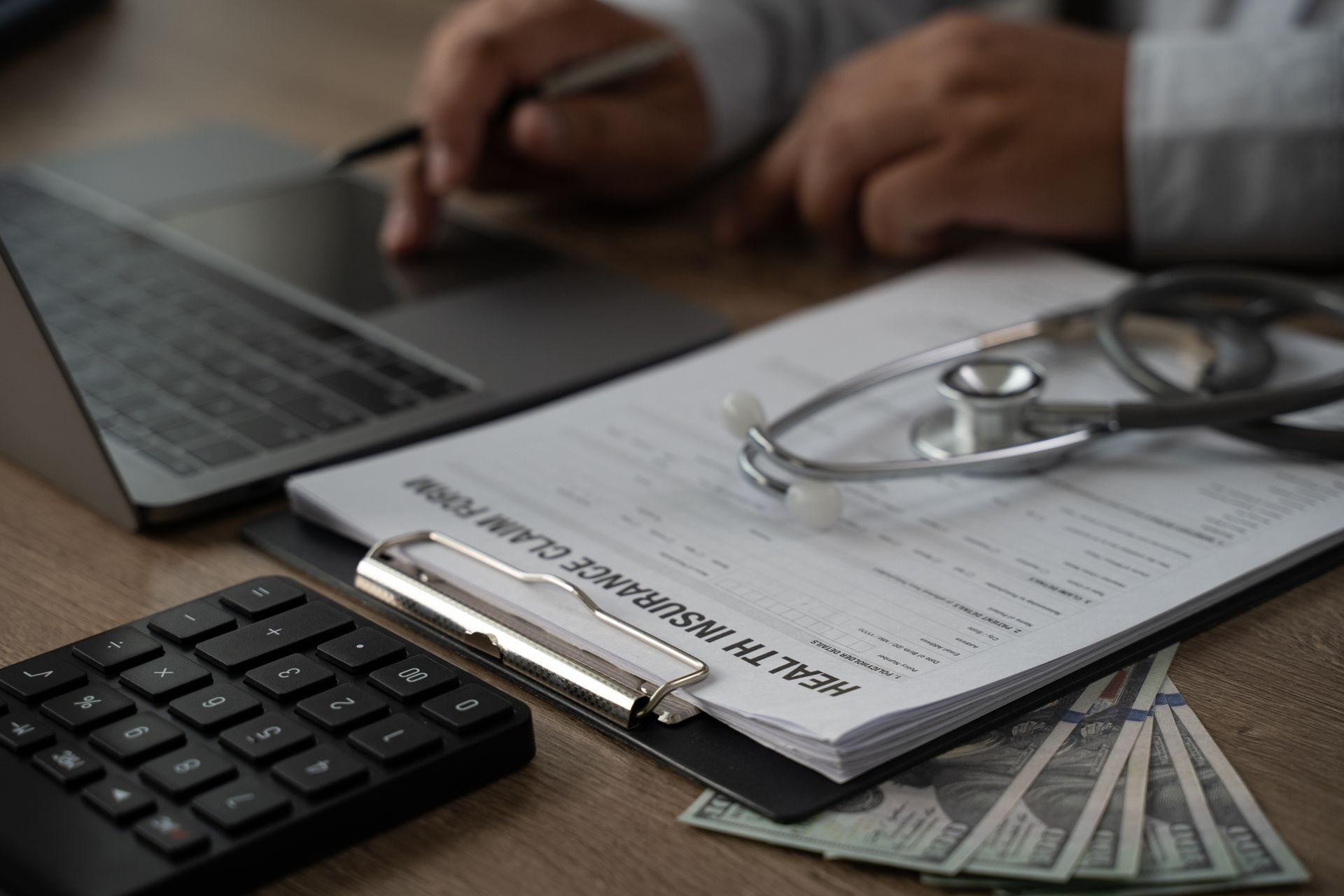 Clipboard with financial paperwork beside a laptop, calculator, stethoscope, and a hand writing notes
