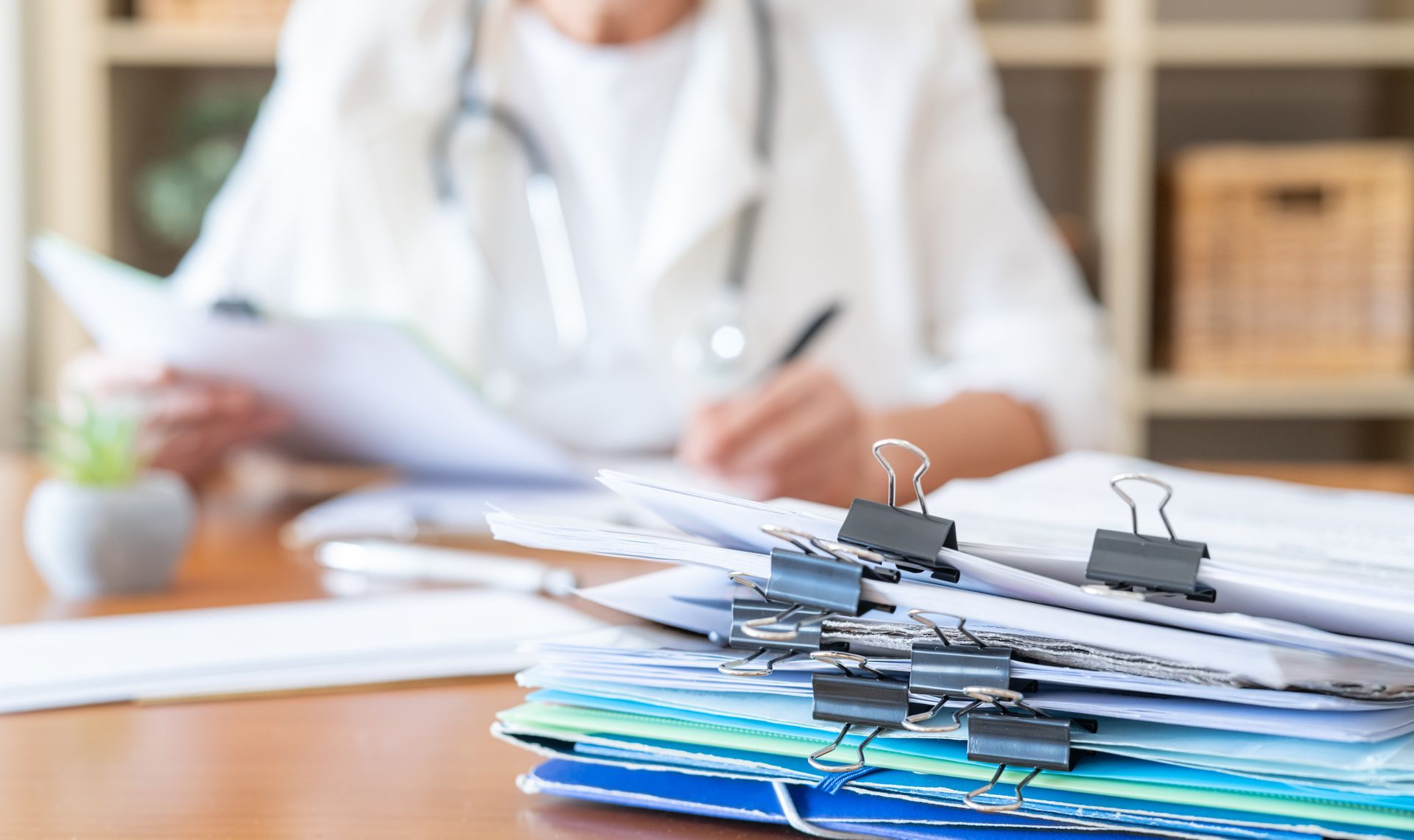 Stack of medical records and folders on a desk with a doctor writing in the background