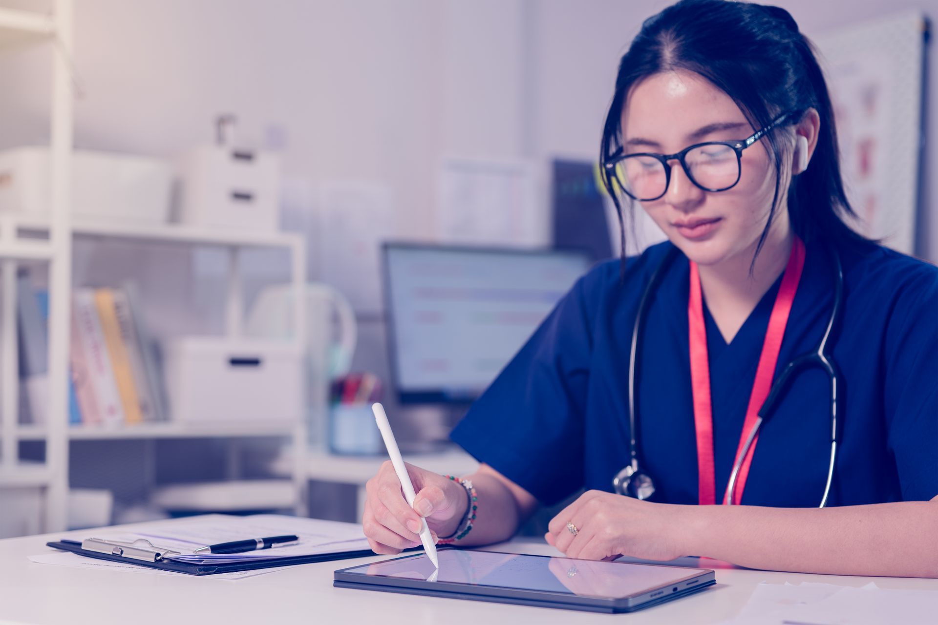 Woman writing in a notebook at a desk, wearing glasses and a red lanyard in a bright office