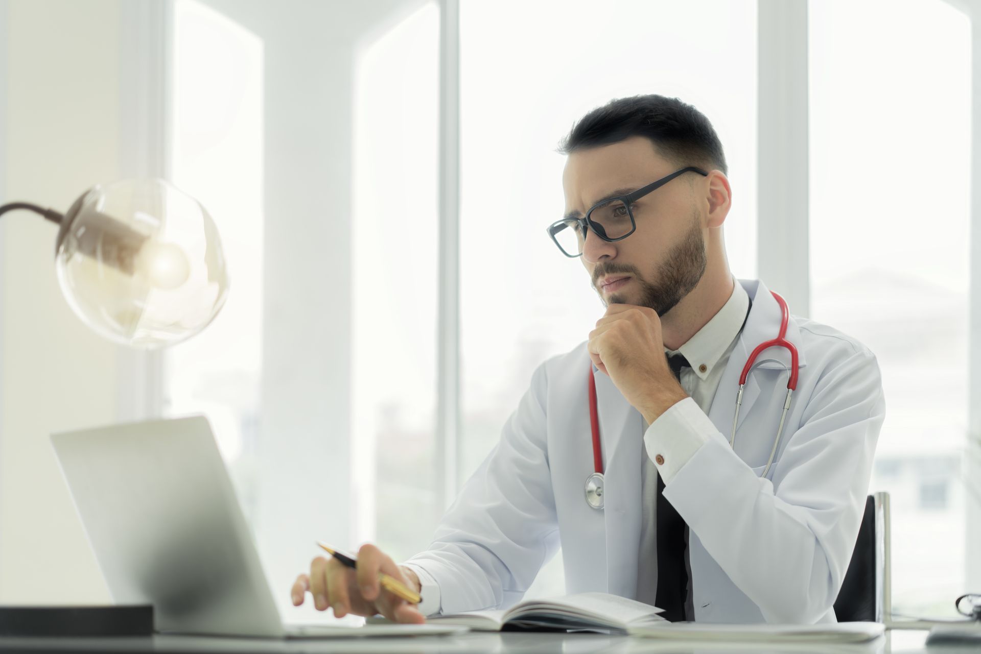 Doctor in a white coat, resting his chin while reviewing a laptop at a bright office desk.
