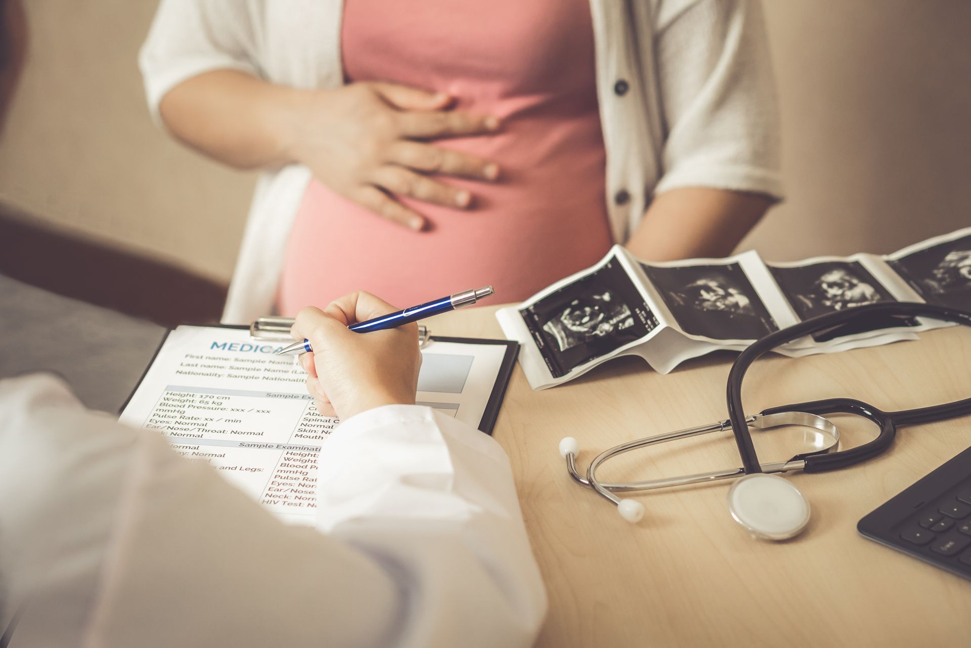 Doctor consulting with pregnant patient; ultrasound images and stethoscope on desk.