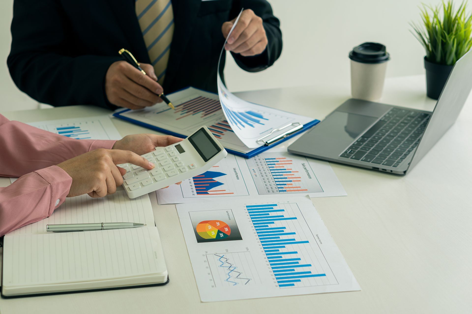 Two people reviewing charts and a calculator at a desk with a laptop, notebook, and coffee cup