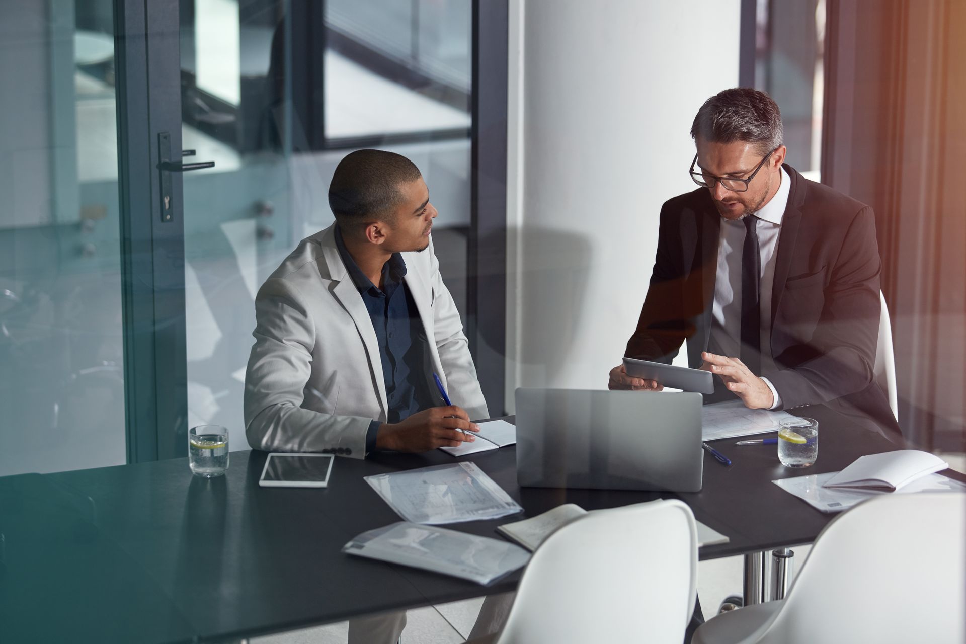 Two colleagues discuss paperwork at a conference table in a modern glass-walled office.