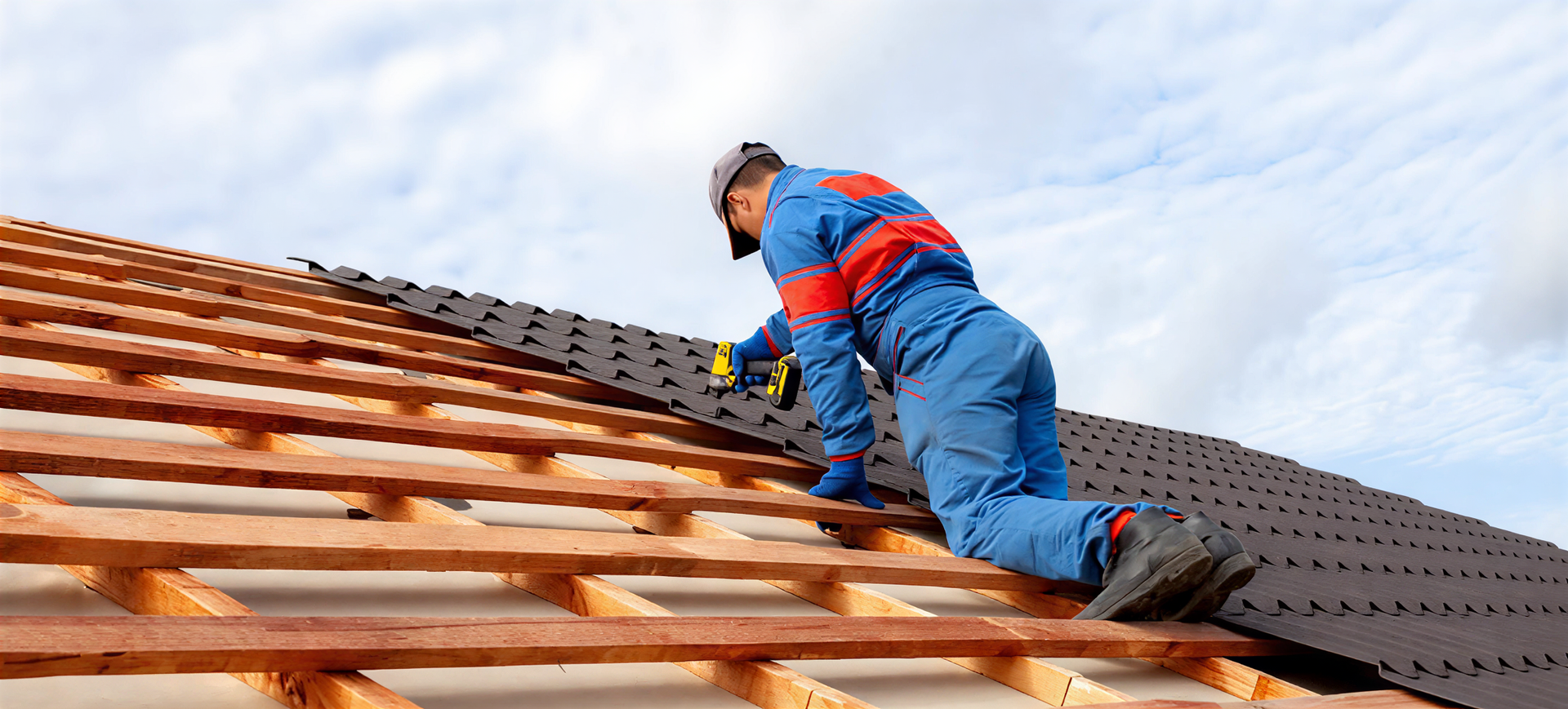 Worker installing roof shingles on a house roof, wearing blue work clothes and a hard hat.