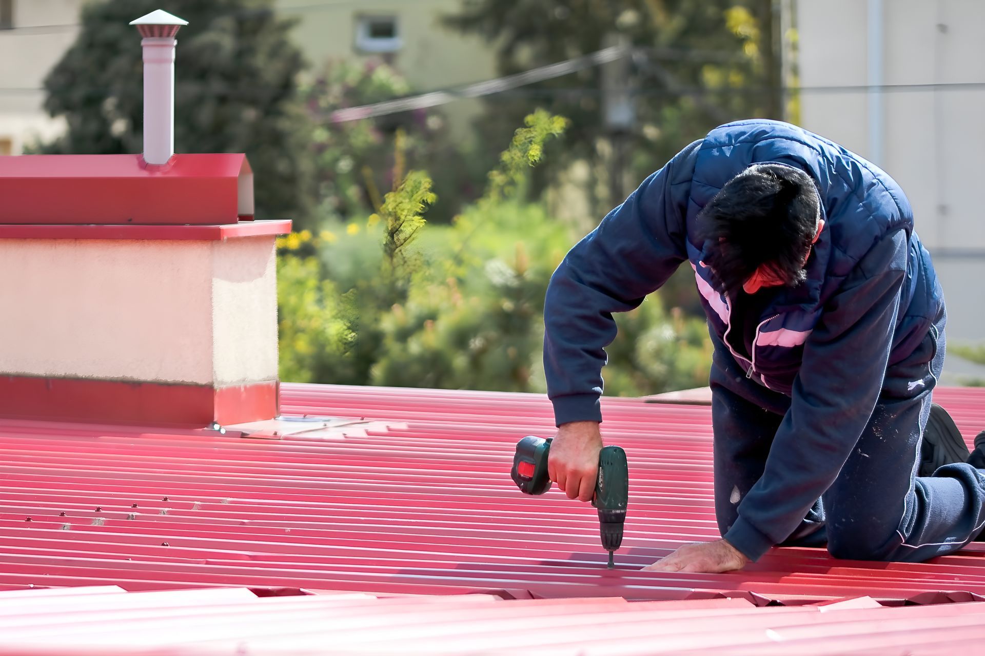 Person kneeling on a red roof, using a drill near a white vent in bright daylight