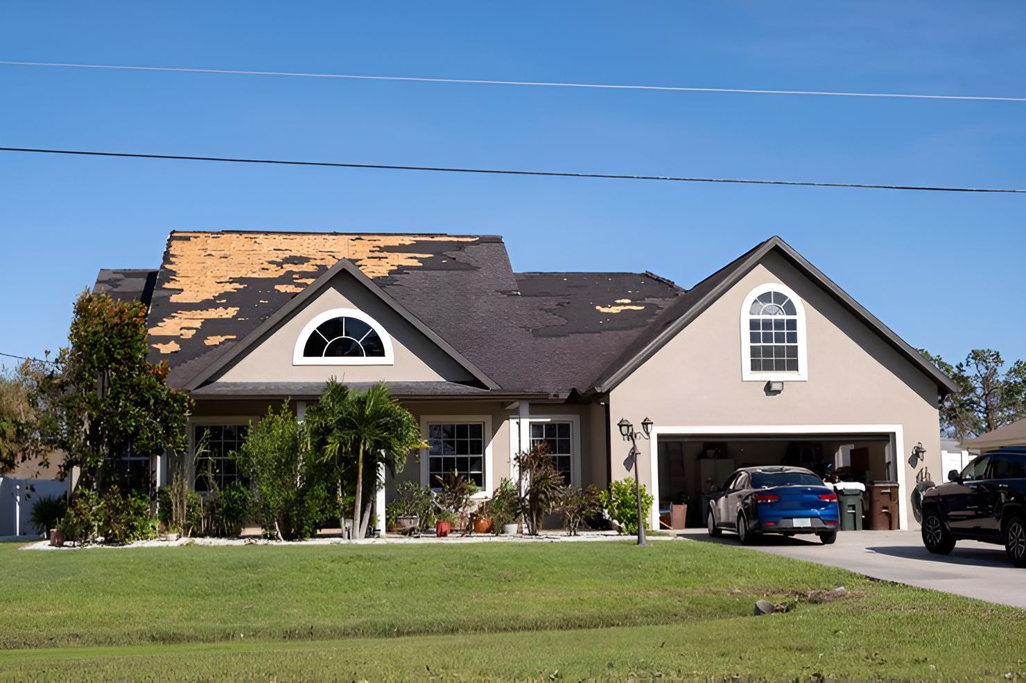 Single-story suburban house with a dark roof, manicured lawn, and two cars in the driveway under a blue sky