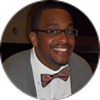 Smiling man wearing glasses, a bow tie, and a gray suit jacket at an indoor event