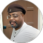 Man in a black beret and white shirt smiling in a doorway, shown in a circular crop
