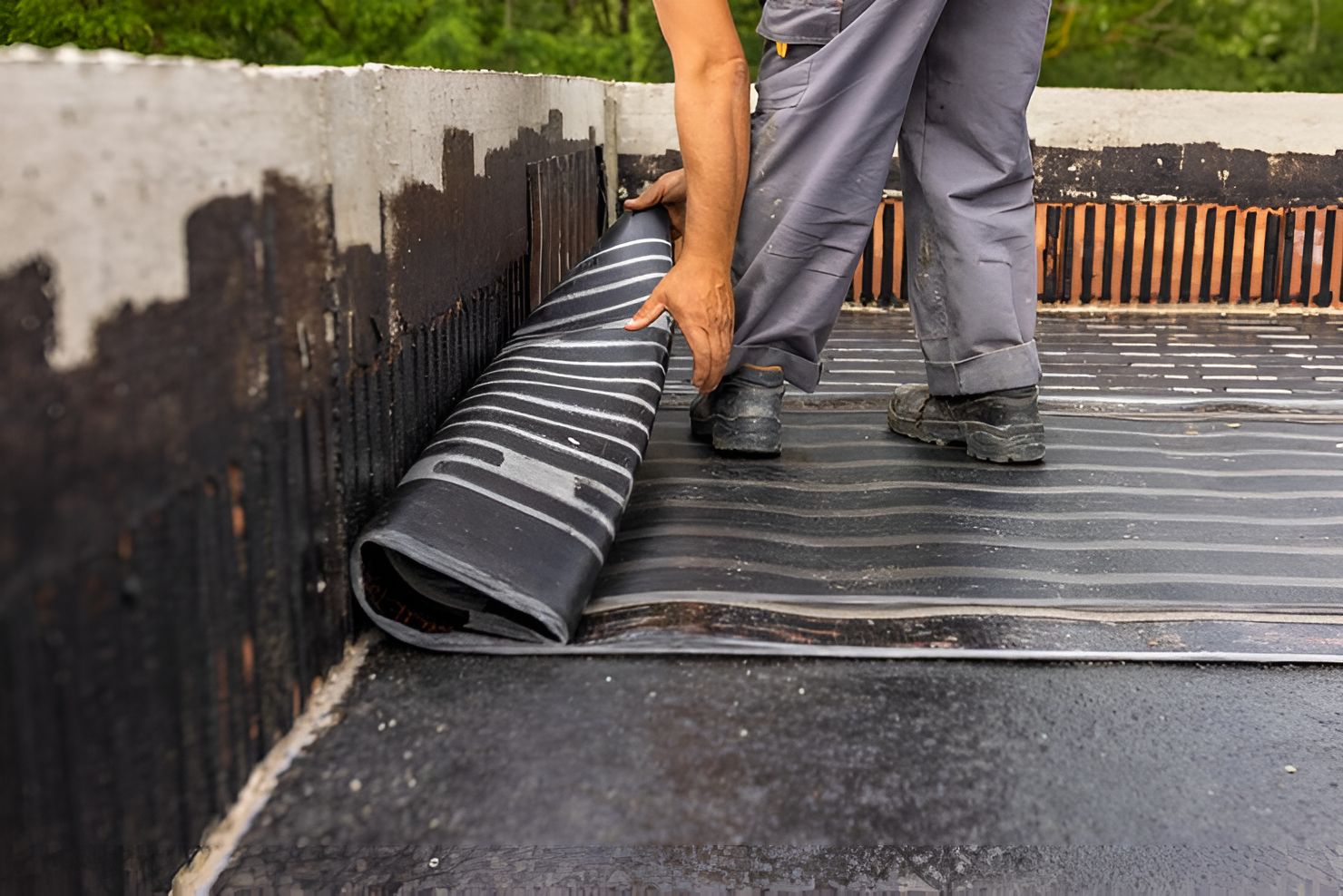 Worker unrolling black roofing membrane on a flat roof alongside a wall