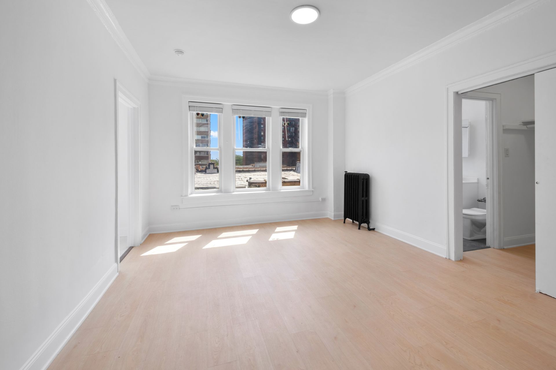 Empty room with light wood floors, large windows, and a doorway to a bathroom.