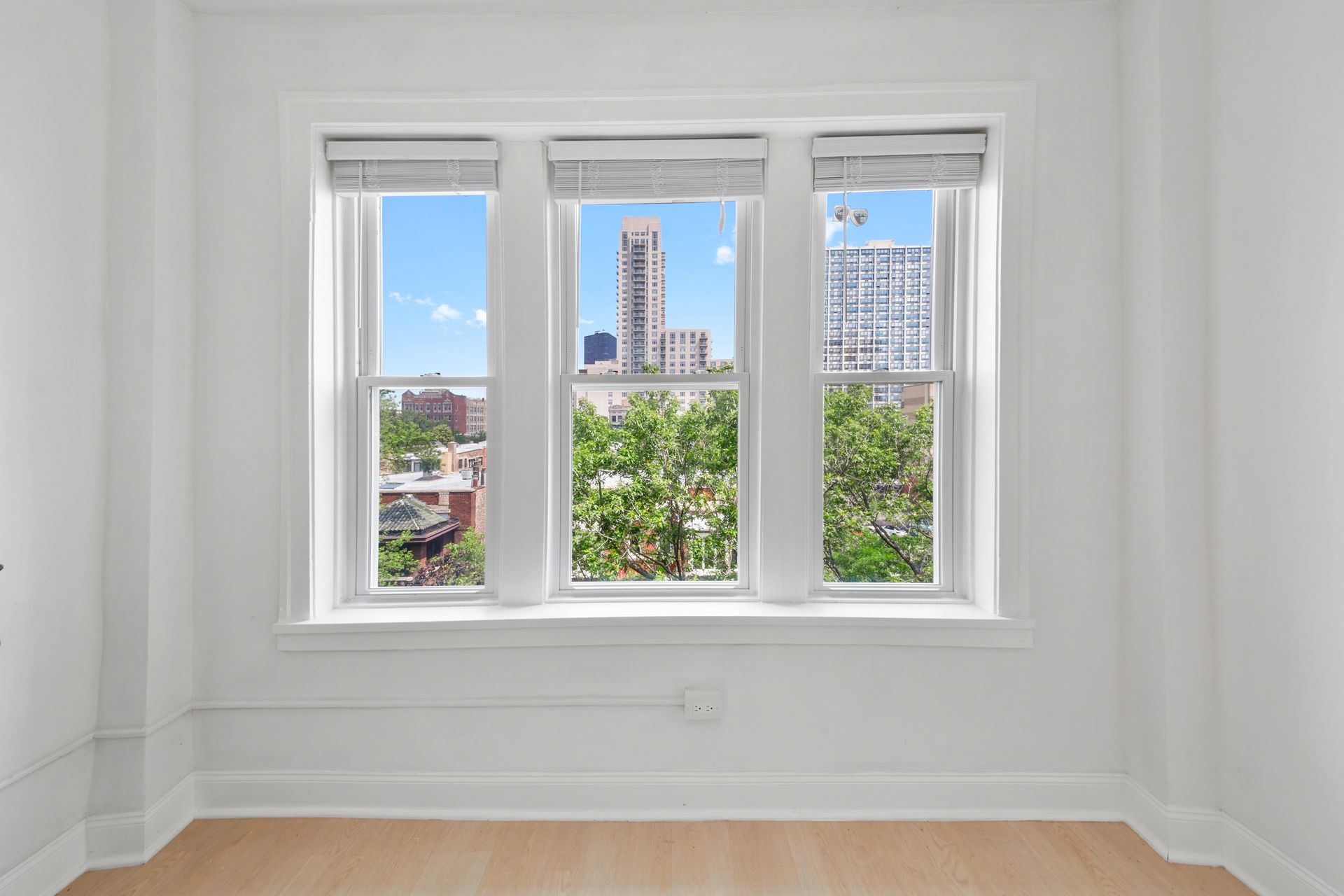 A bright room with a triple-paned window overlooking a city with trees and buildings; white walls, beige floor.