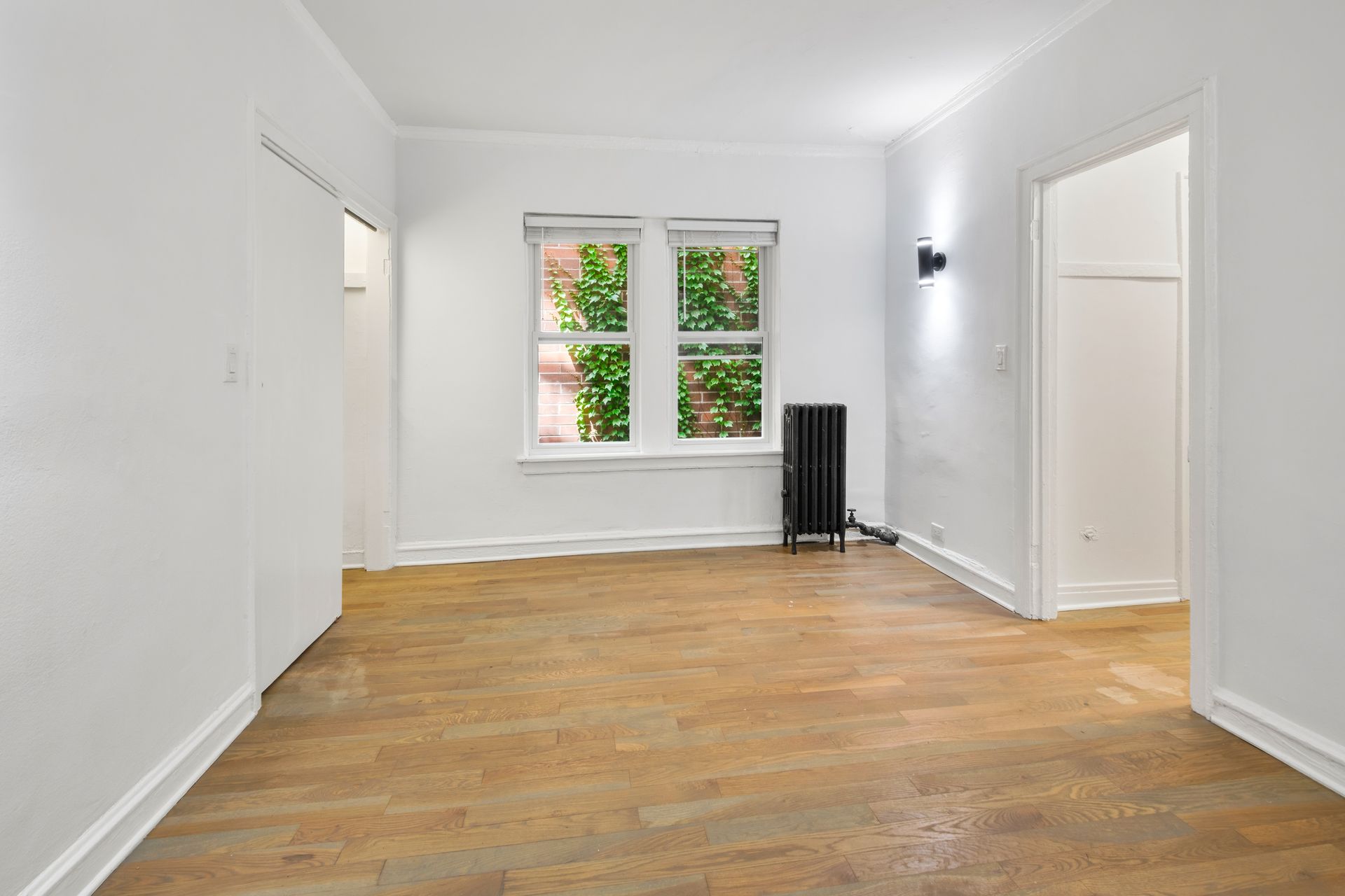 Empty room with wooden floor, window with greenery, and an open doorway.
