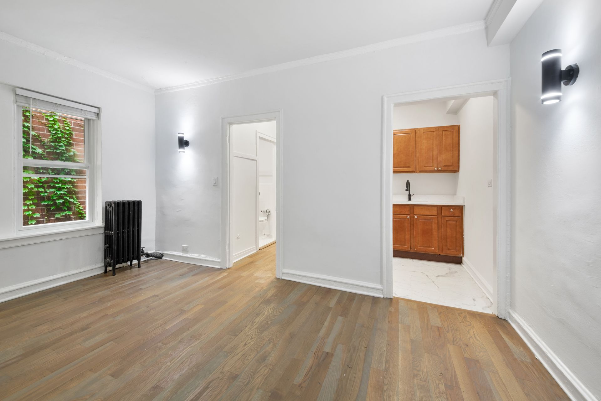 Empty, white-walled room with hardwood floors, two open doorways, and a window with outdoor foliage.