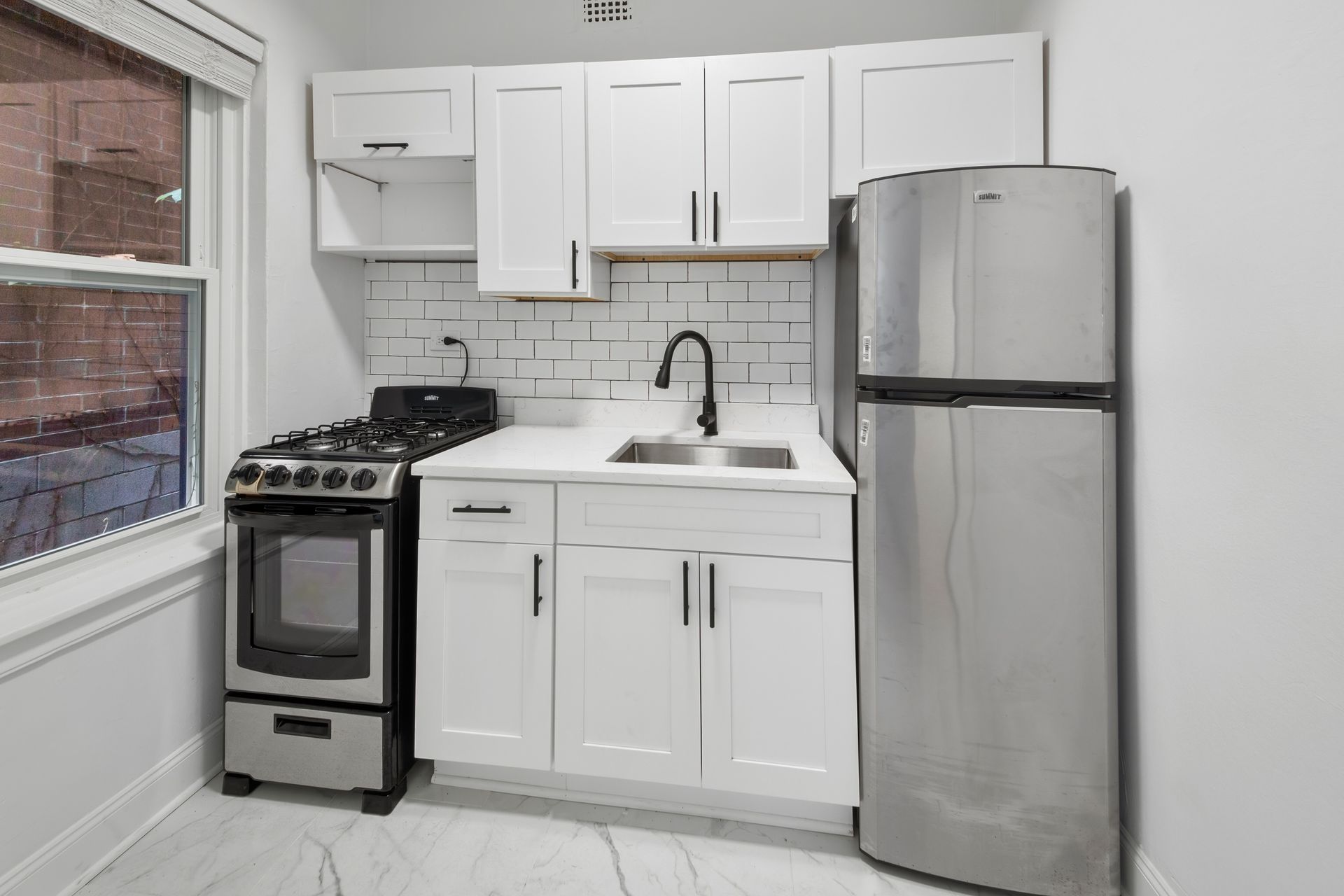 Small, white kitchen with a stove, cabinets, sink, and a stainless steel refrigerator.