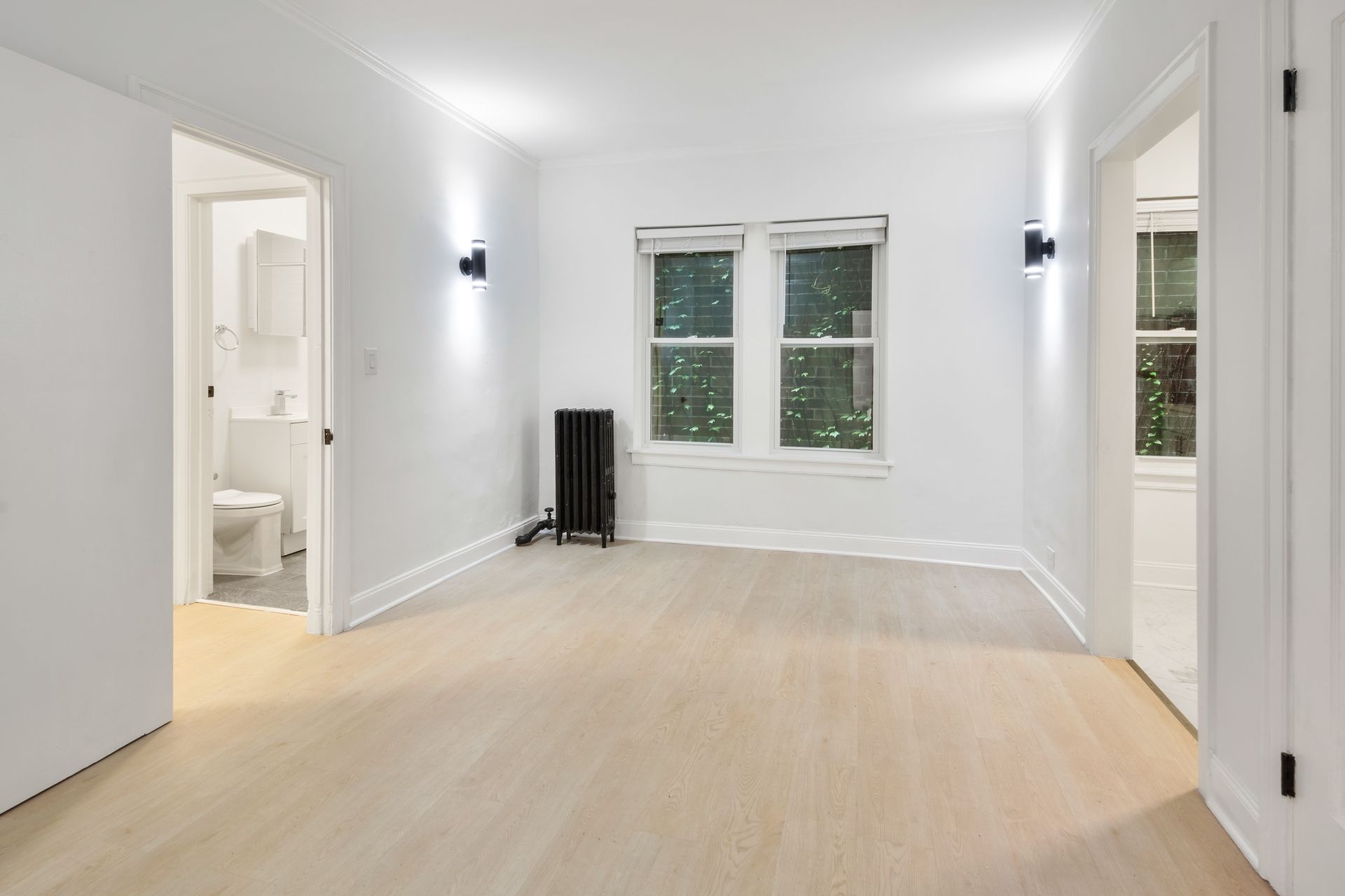 Empty white-walled room with a bathroom and another room visible through doorways. Window and small radiator present.