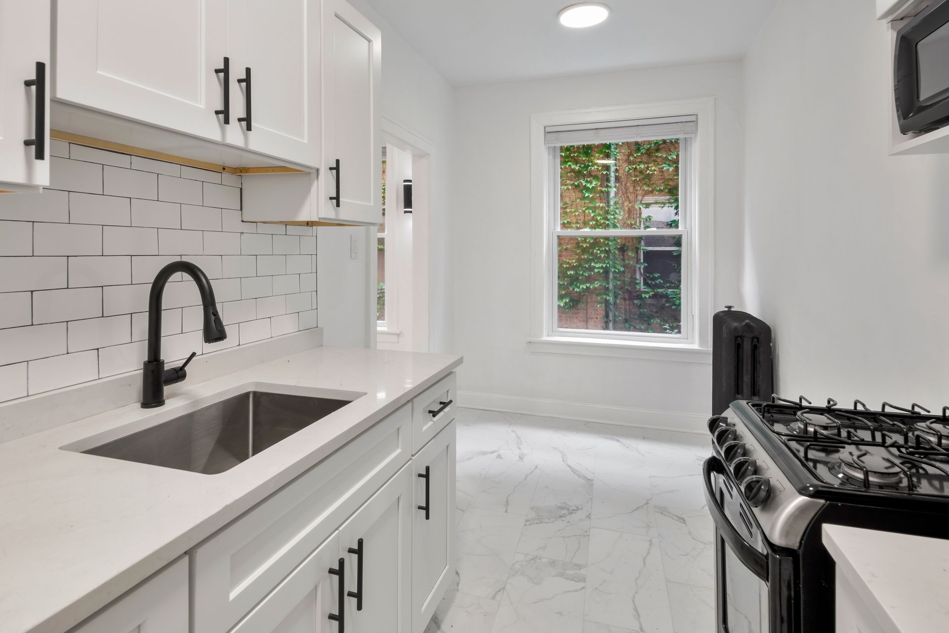 White kitchen with black accents: cabinets, faucet, handles. Window, stove, sink, white tile backsplash.
