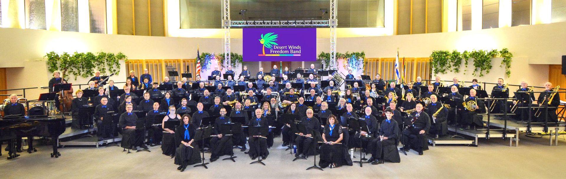 Photo of the symphonic band from the 2025 spring concert at Temple Isaiah/Jewish Community Center.  The band is dressed in black with blue ties and scarves and are sitting on risers on the stage at the venue.