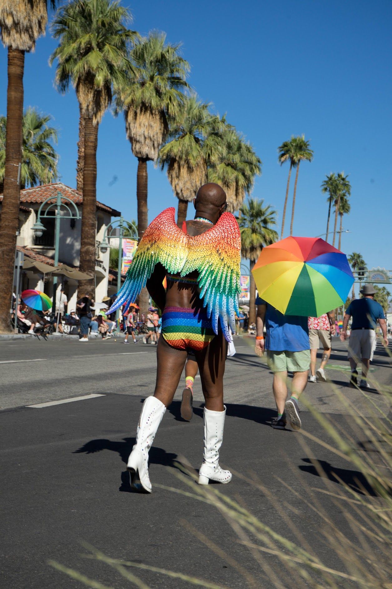 Phtoto from the 2026 Pride Celebration in Palm Springs showing a man walking in rainbow shorts and wearing rainbow angel wings. Others are carrying rainbow colored sun umbrellas.