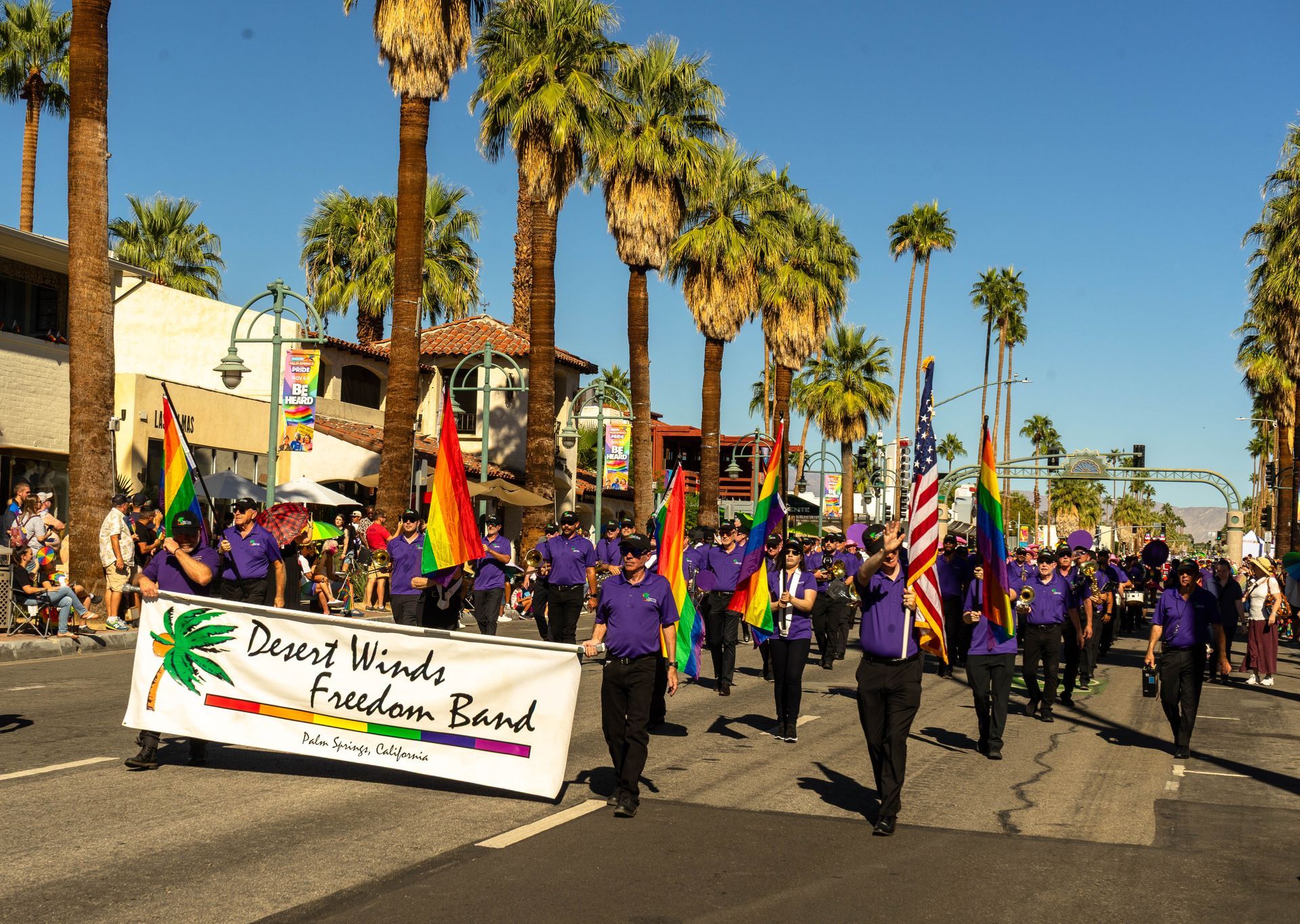 Photo of the marching band at the 2025 Pride Parade in Palm Springs.  The band is dressed in black pants and purple band shirts.  There is a color guard with rainbow flags.  A crowd 5 deep lines the sidewalks.