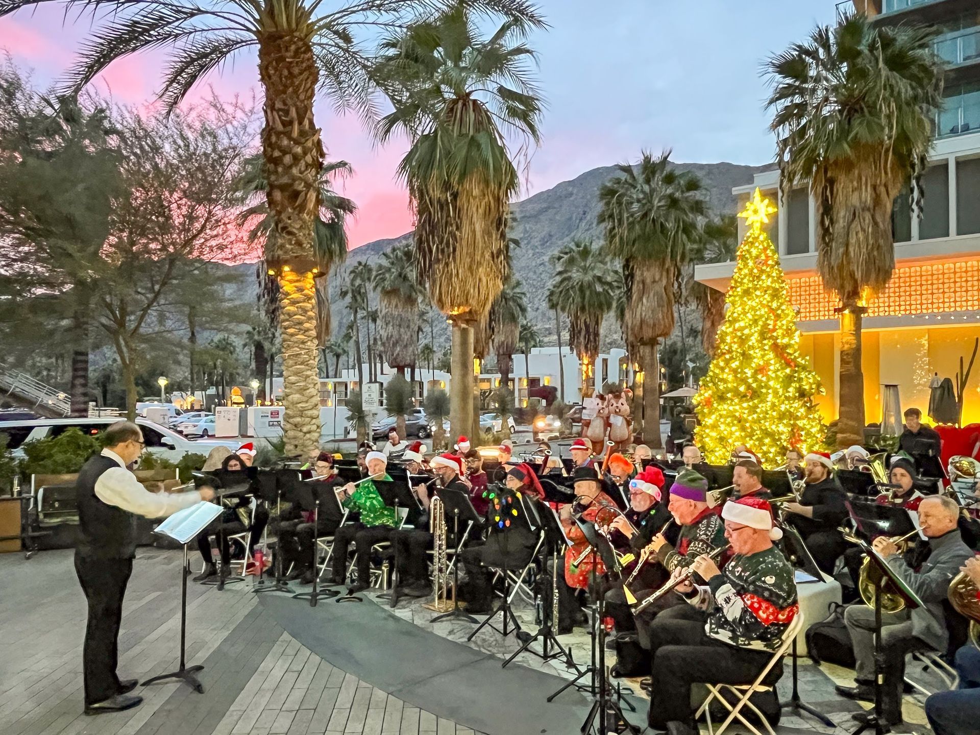 The Pep Band is shown here playing a concert of Holiday music in downtown Palm Springs on the plaza near Starbucks.  In the background are mountains, palm trees and Christmas decorations.  The band is dressed un ugly Christmas sweaters and many are wearing Santa hats or flashing lights.