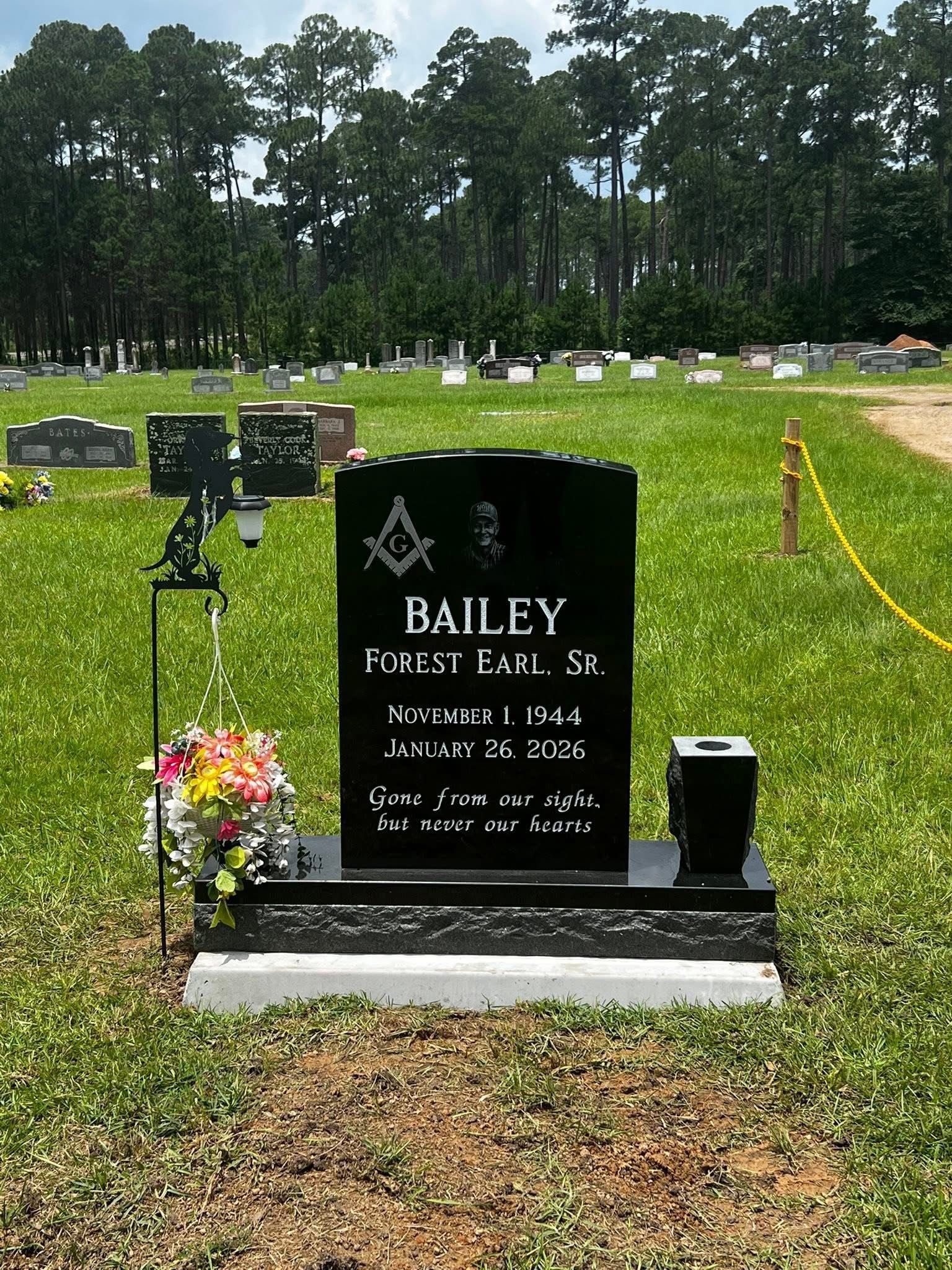 Black Bailey gravestone with flowers in a cemetery, green lawn and trees in the background.