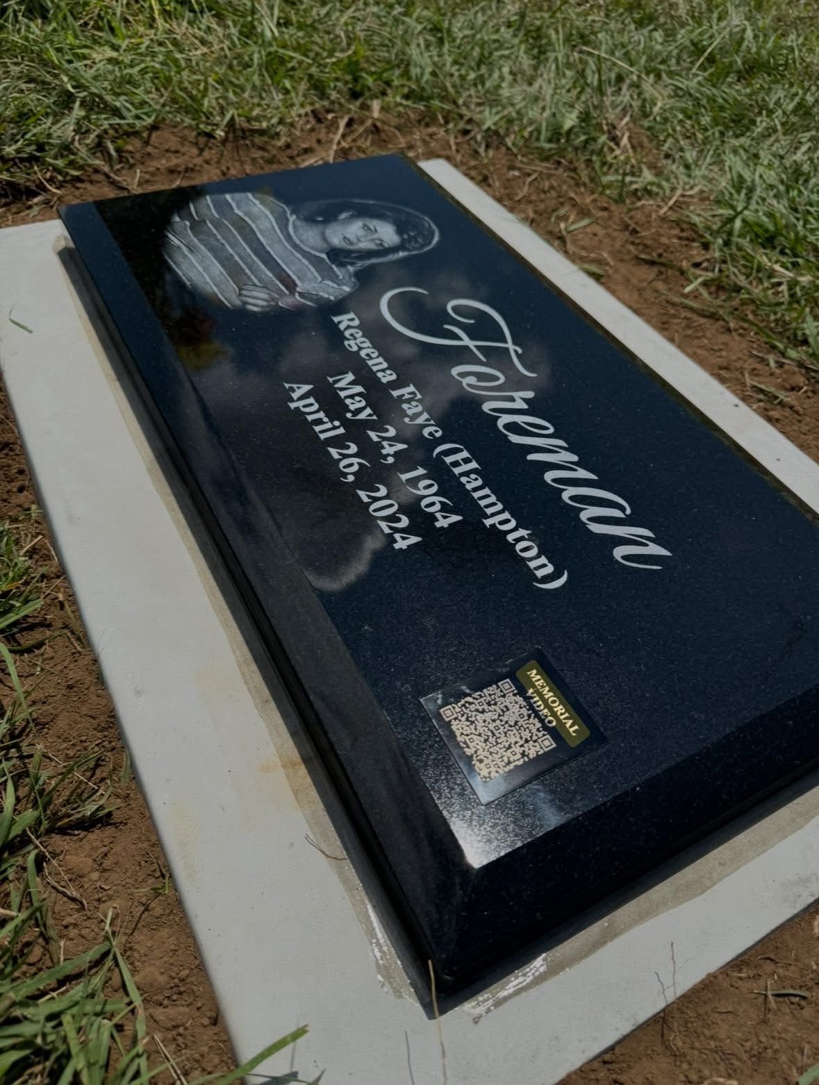 Black memorial grave marker on a concrete base in grass, with engraved portrait and white lettering.