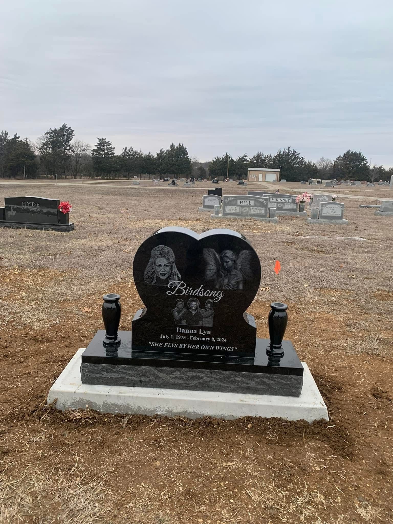 Black heart-shaped memorial headstone in a cemetery with a gravel landscape and cloudy sky.